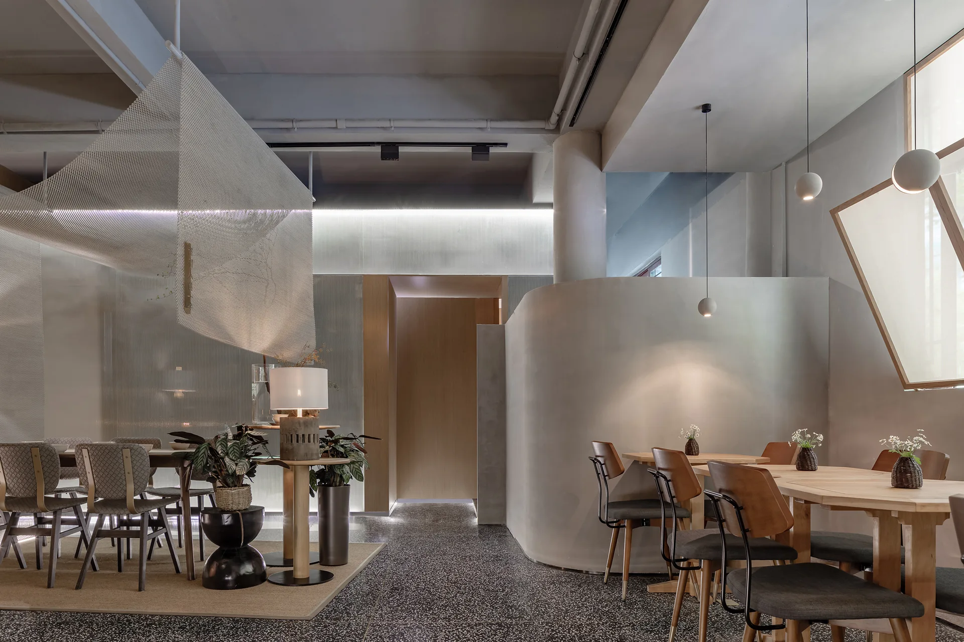 A contemporary dining room featuring wooden tables and chairs, a smooth curved partition wall, and a hanging white metal mesh installation.