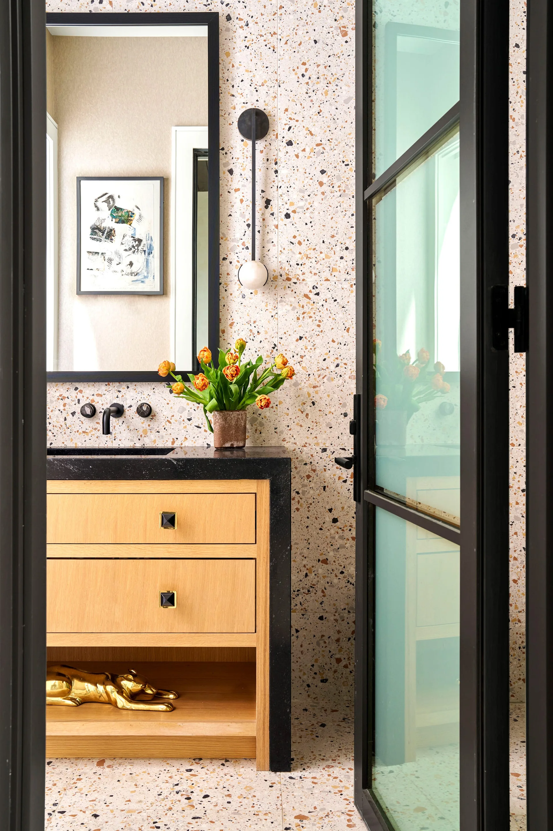 A modern powder room features a light wood vanity with a black countertop set against a speckled terrazzo tile wall and a black framed mirror.