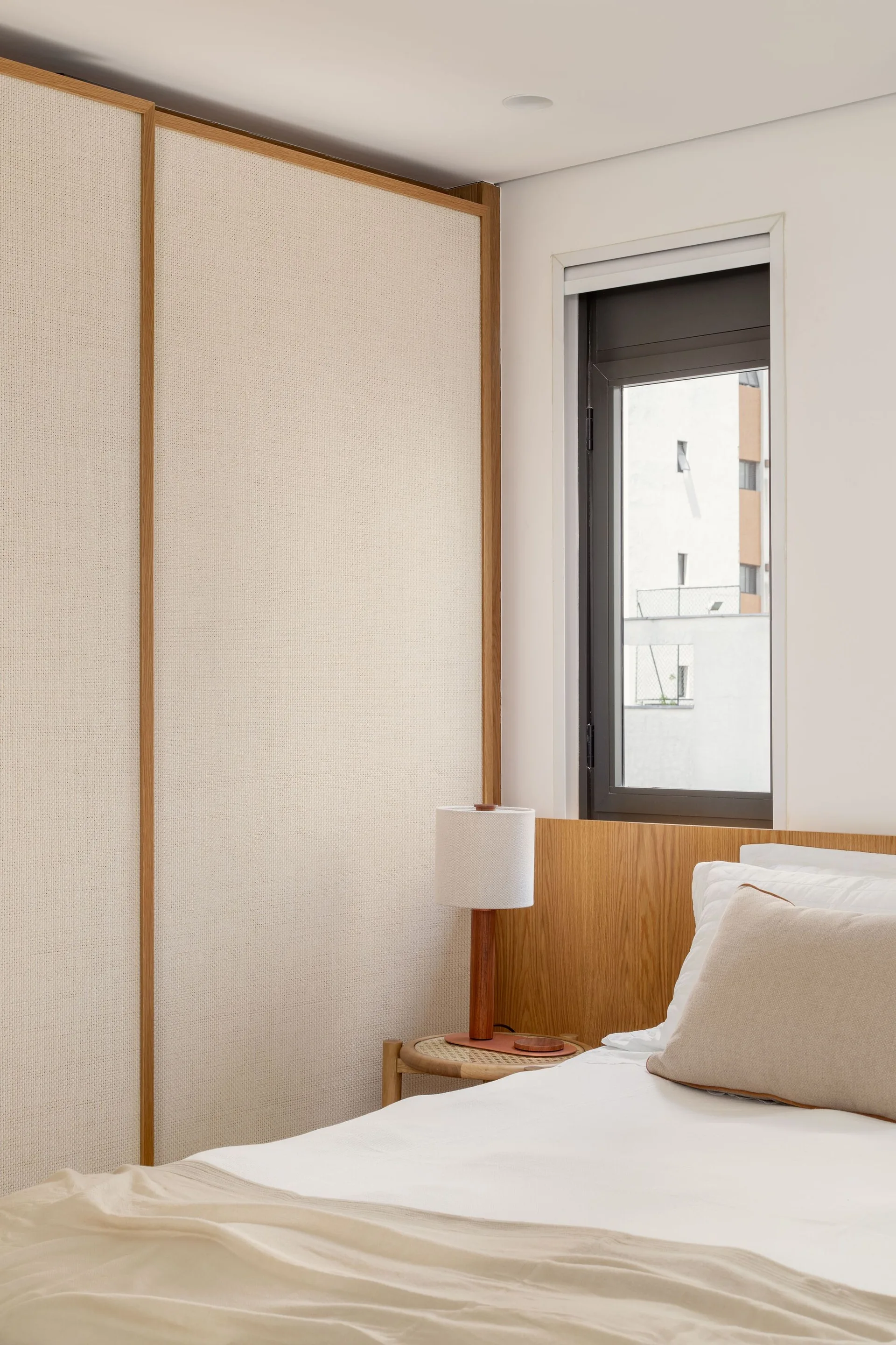 A minimalist bedroom corner featuring a sliding wardrobe with woven fabric panels next to a low wood headboard and a round rattan nightstand.