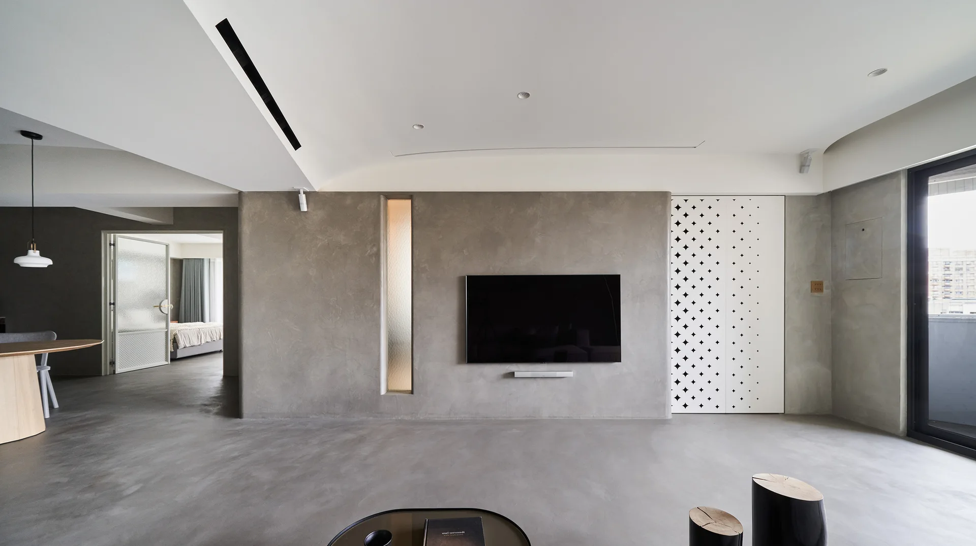 A minimalist living room features smooth grey concrete walls and flooring, a wall-mounted television, and a white sliding door with star-shaped perforations.