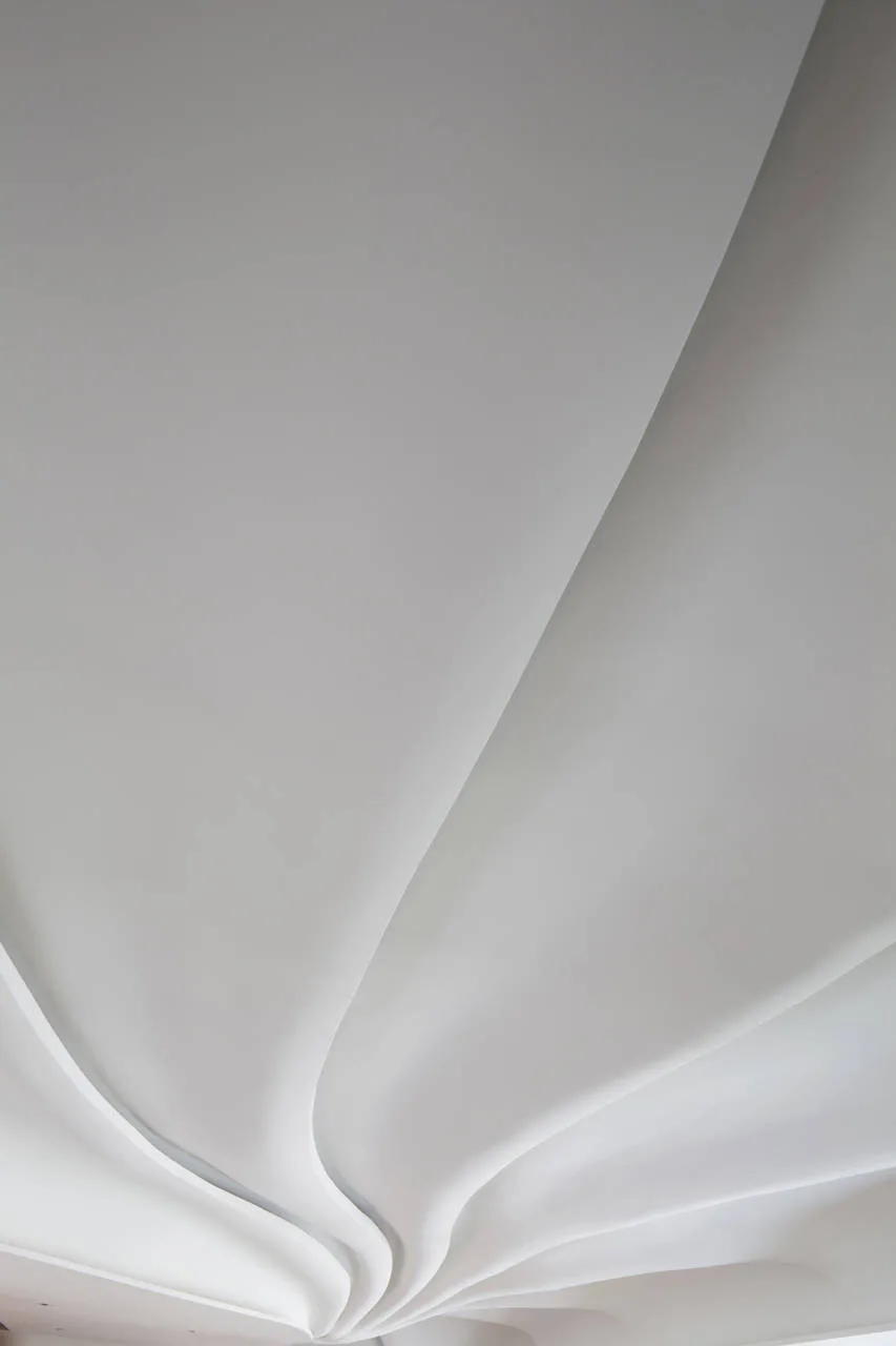 A close-up view of a minimalist white ceiling featuring smooth, fluid, sculptural curves and soft shadows on its matte surface.