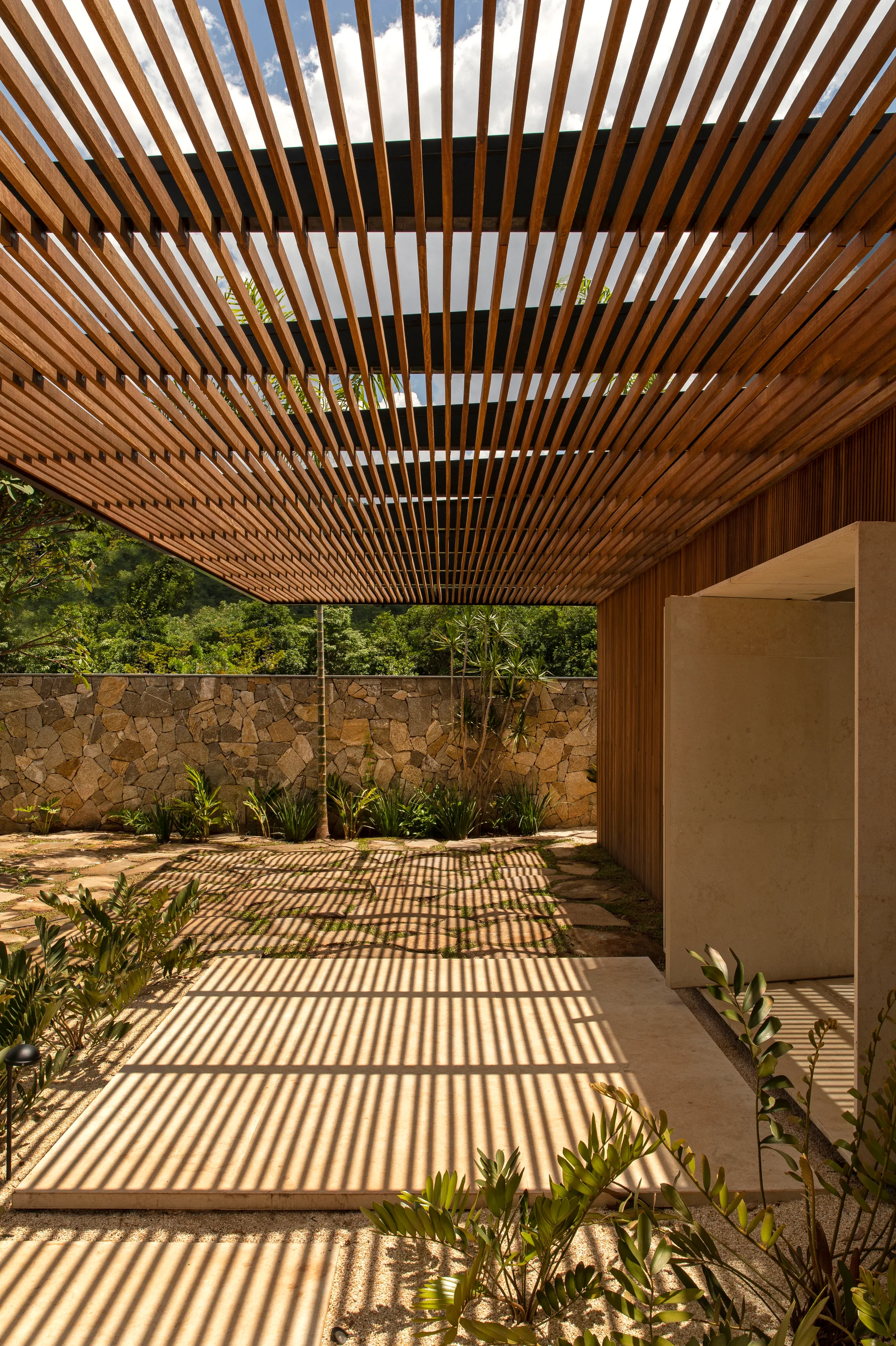 A wooden slatted pergola casts linear shadow stripes across a stone-paved garden patio with a rough stone wall in the background.