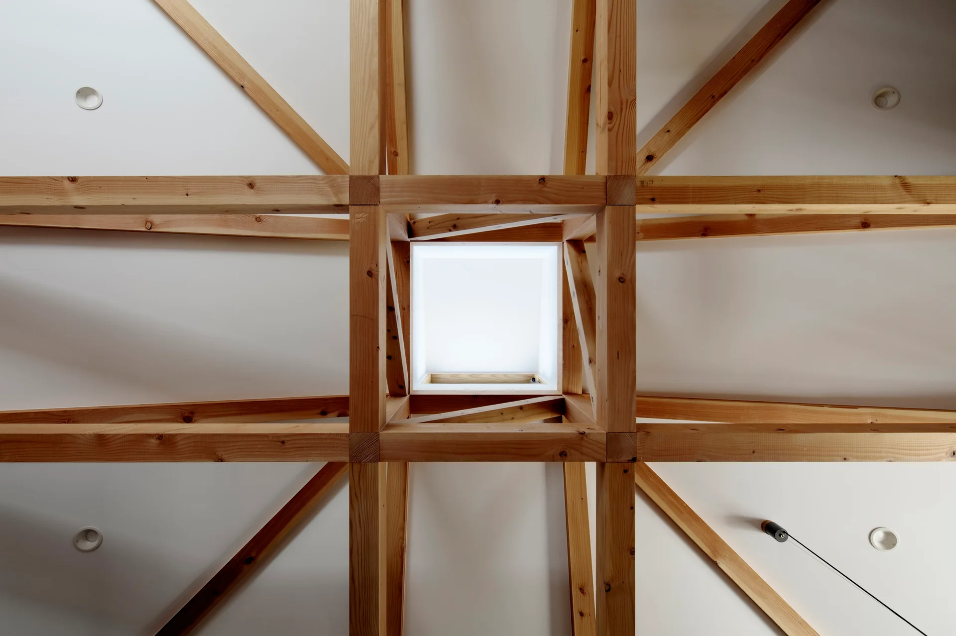 A symmetrical grid of natural wood trusses on a white ceiling surrounds a square central skylight.
