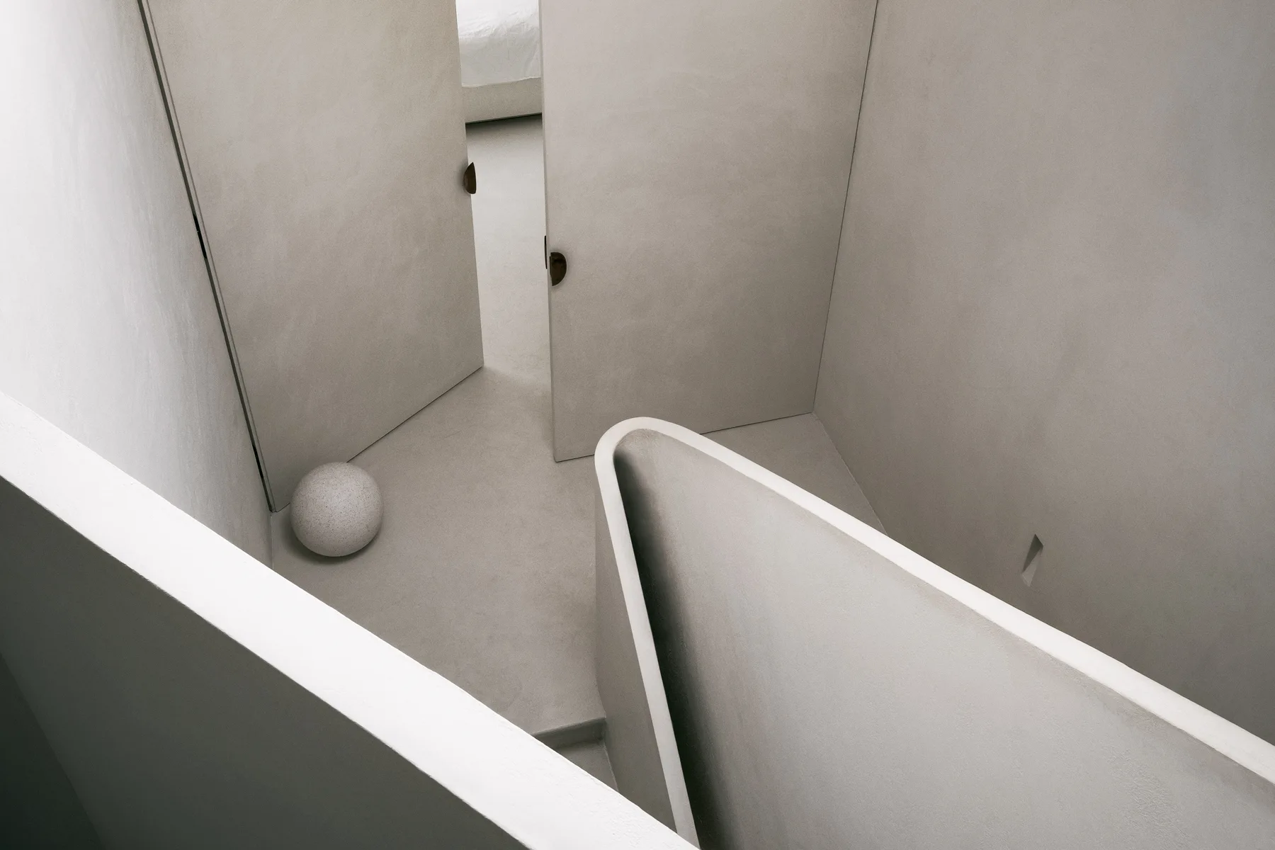 A high-angle view of a minimalist plaster hallway with a curved staircase balustrade, flush doors, and a small stone sphere on the floor.