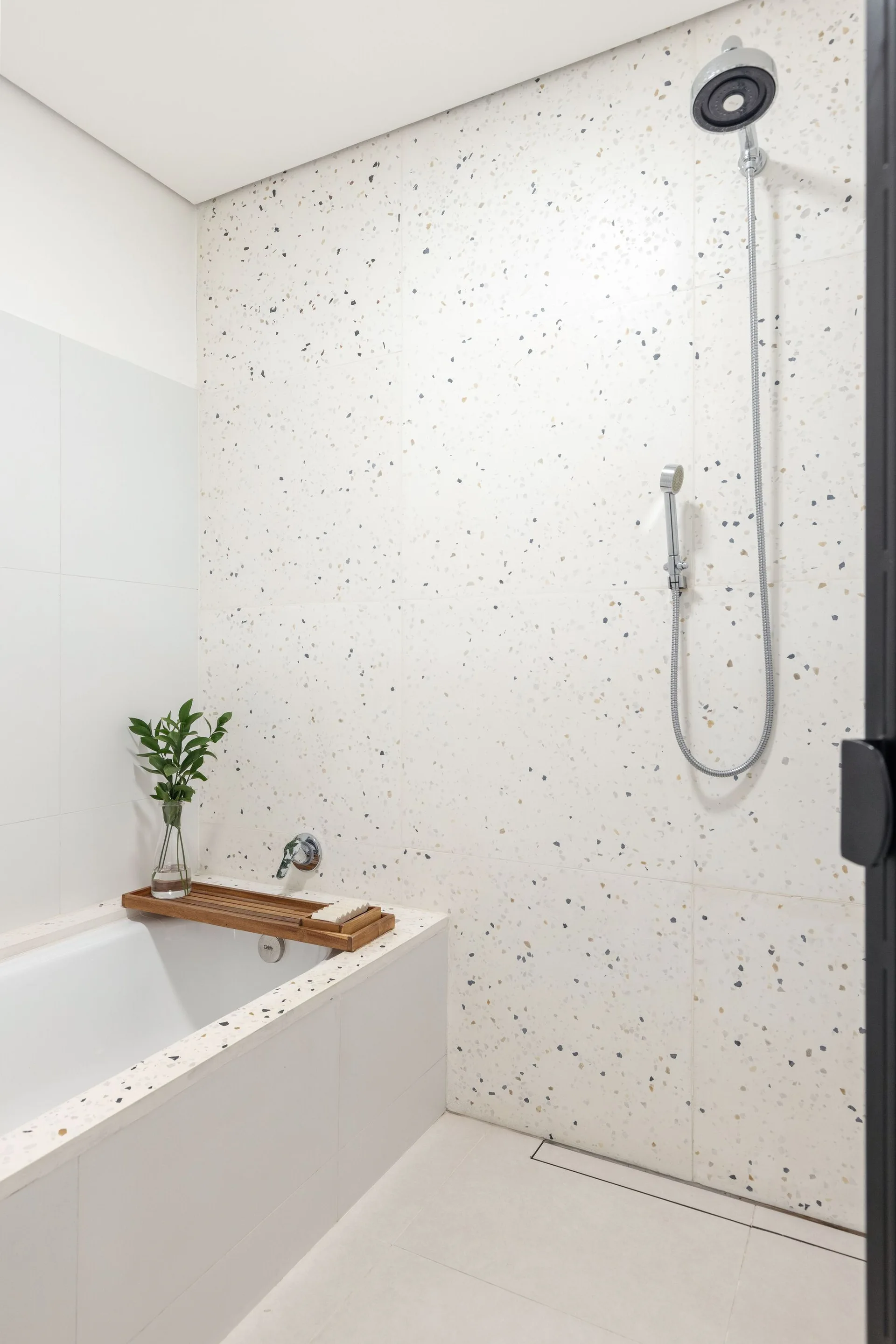 A modern bathroom featuring a white terrazzo tiled wall, a built-in bathtub with a wooden caddy tray, and a wall-mounted chrome shower.