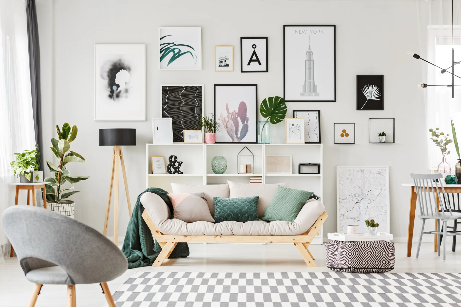 A bright living room features a wood-frame sofa, a white multi-compartment shelving unit topped with a gallery wall of art, and a checkered rug.