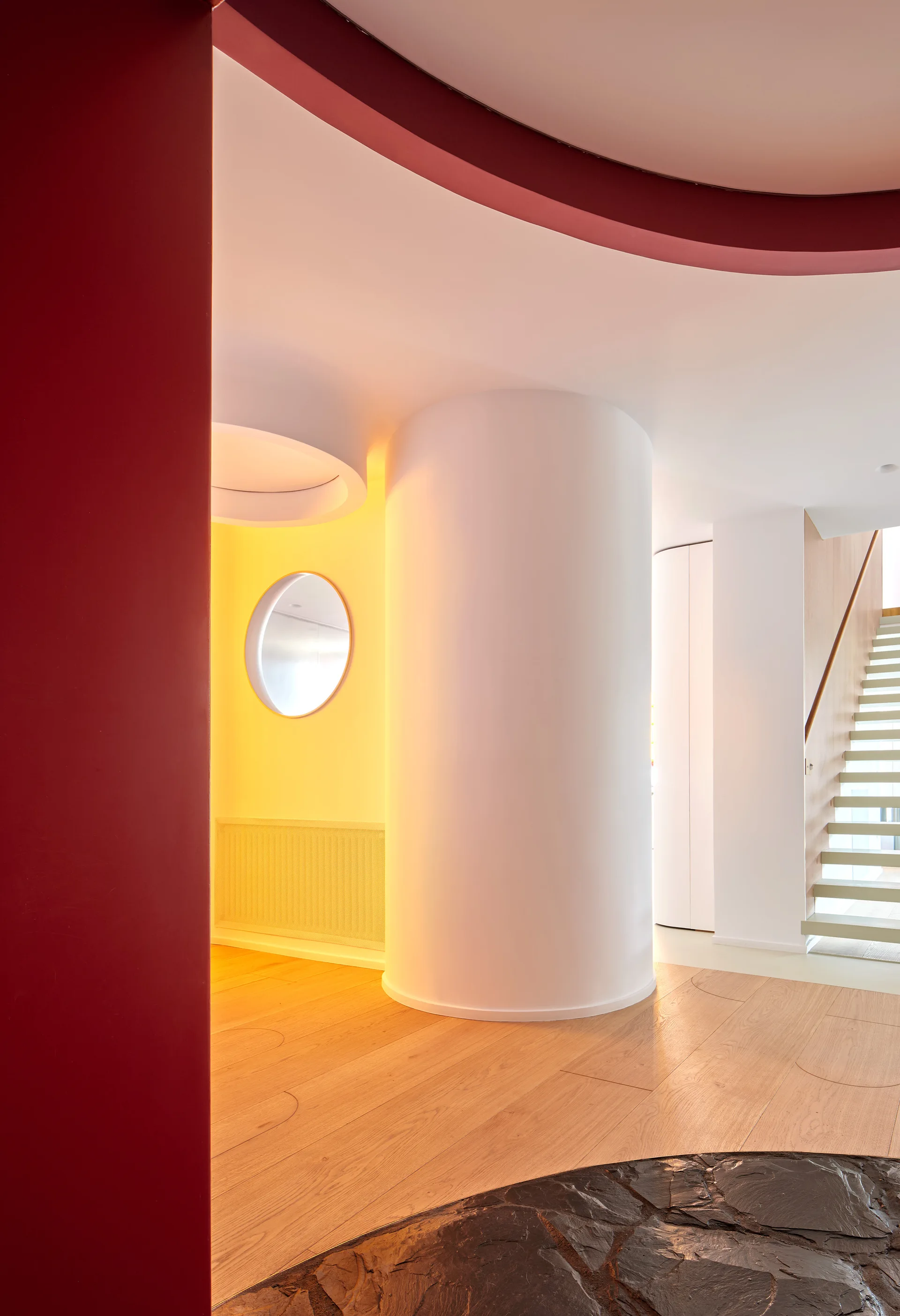 A contemporary hallway features a prominent white cylindrical column, a bold red wall, light wood flooring, and a floating staircase in the background.