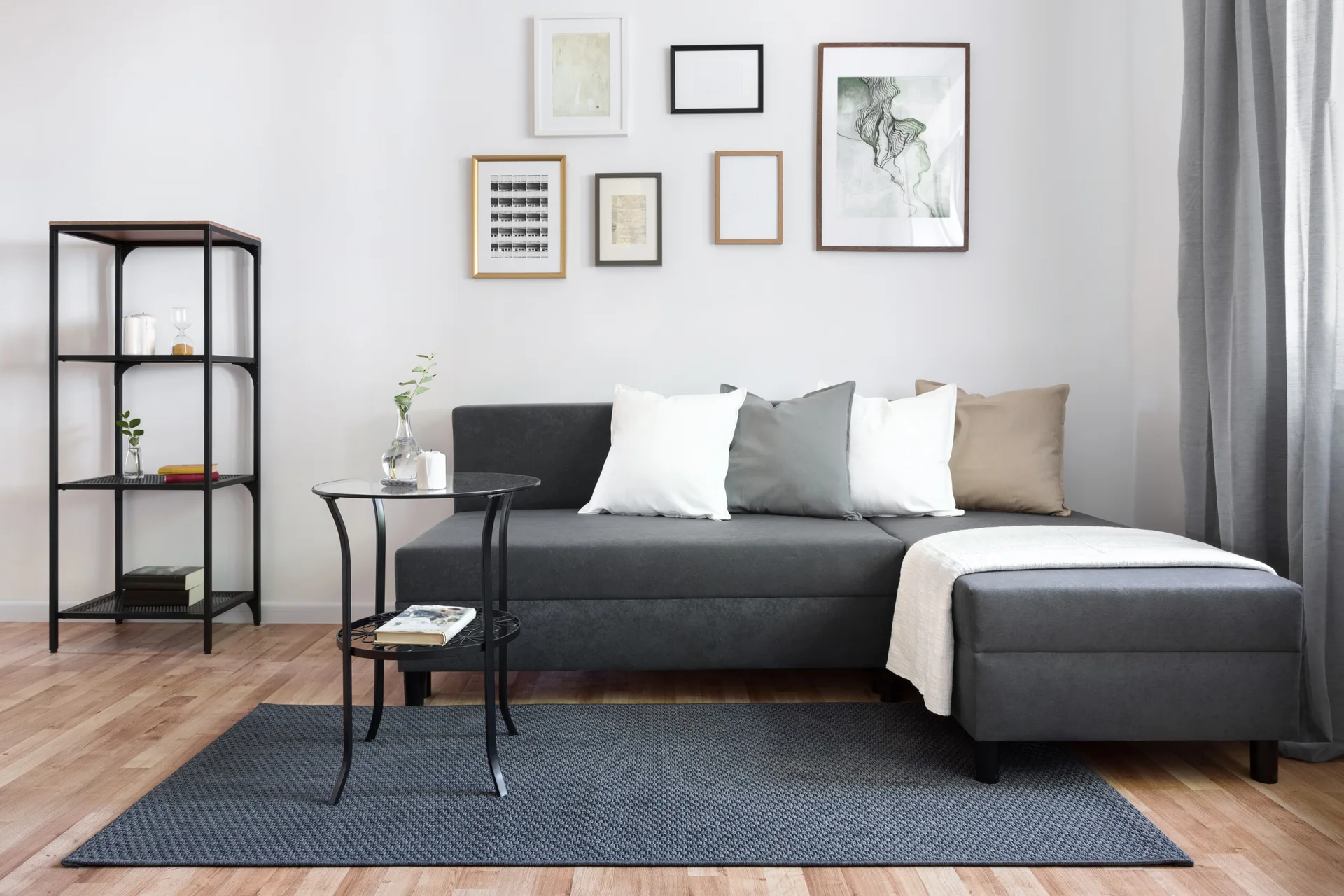 A charcoal grey sectional sofa sits on a dark woven rug next to an open metal shelving unit and a glass-top side table.