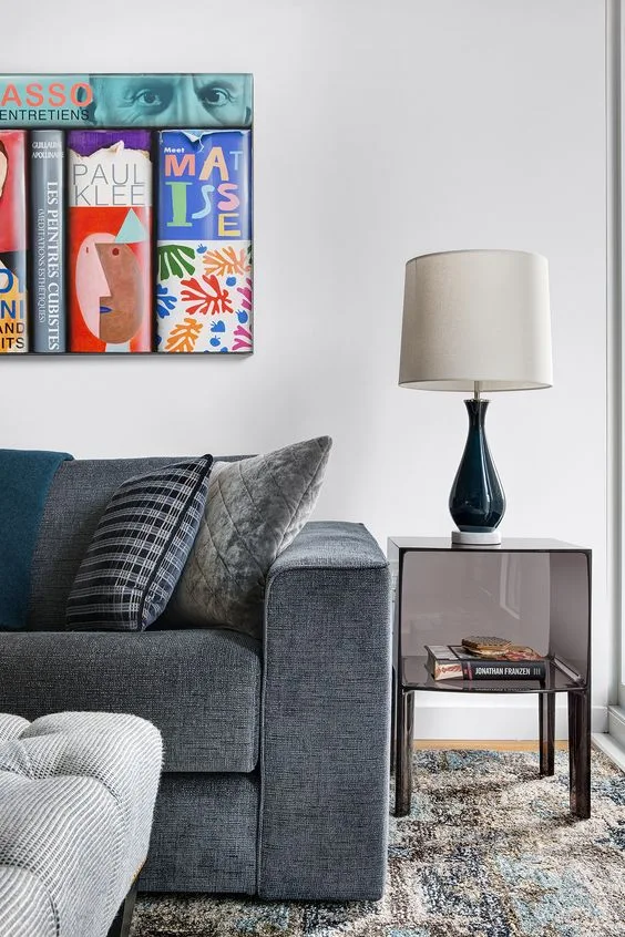 A grey upholstered sofa with plaid pillows sits next to a smoky acrylic side table topped with a blue ceramic lamp in front of a white wall.