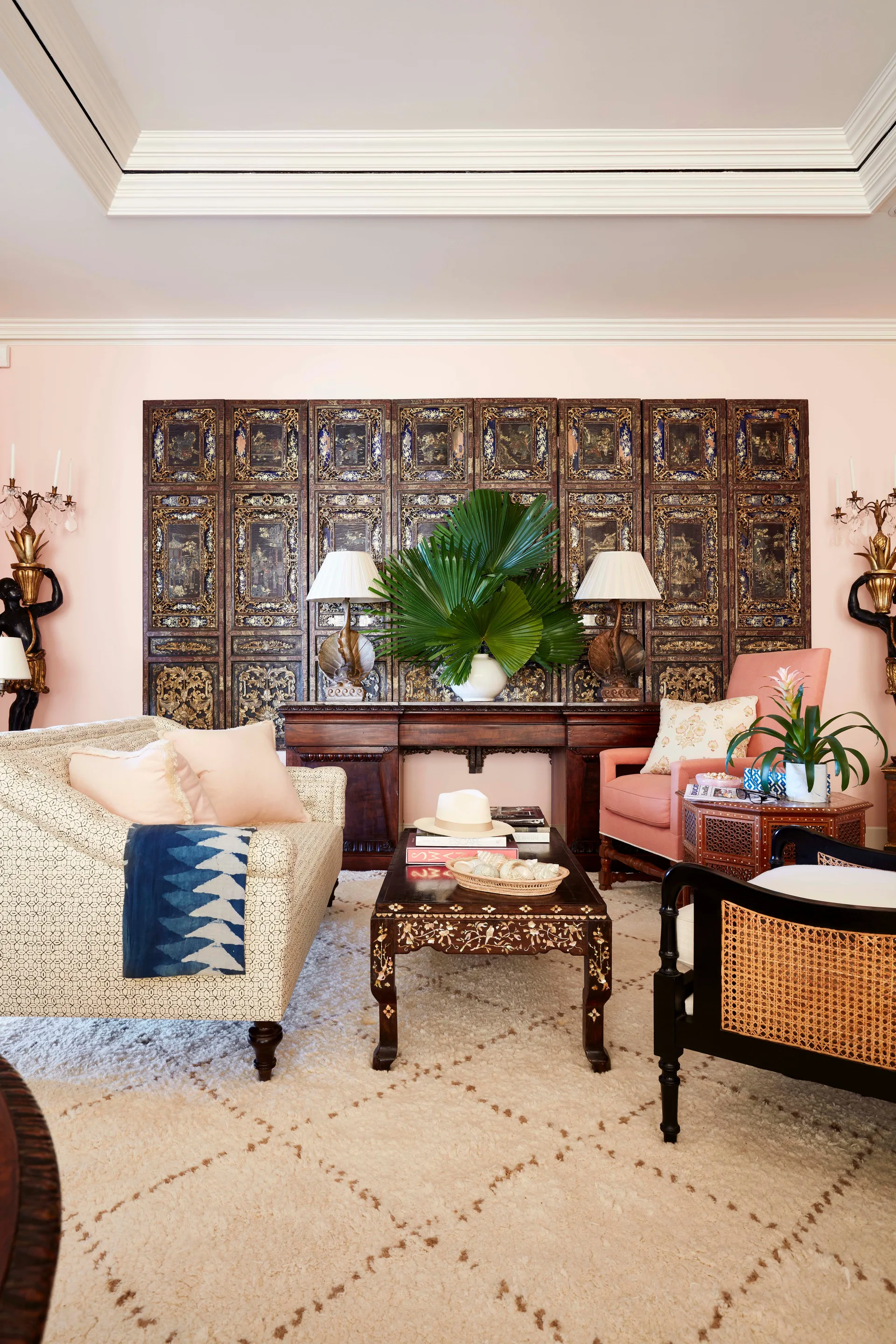 An eclectic living room features a dark wood folding screen behind a console table, a patterned sofa, blush pink walls, and a shag rug.