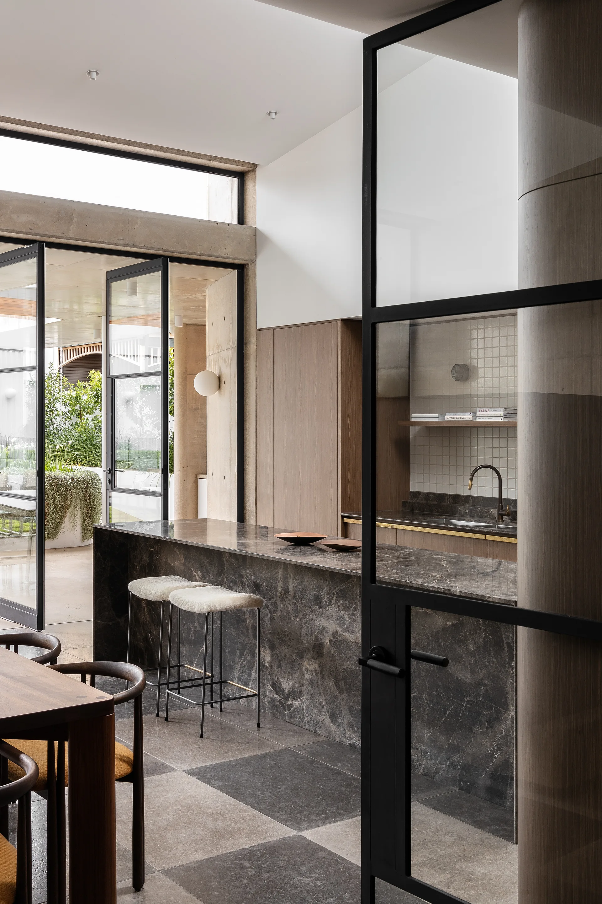 A modern kitchen with a dark marble-topped island, bar stools, and black steel-framed glass doors opening to a concrete outdoor patio.