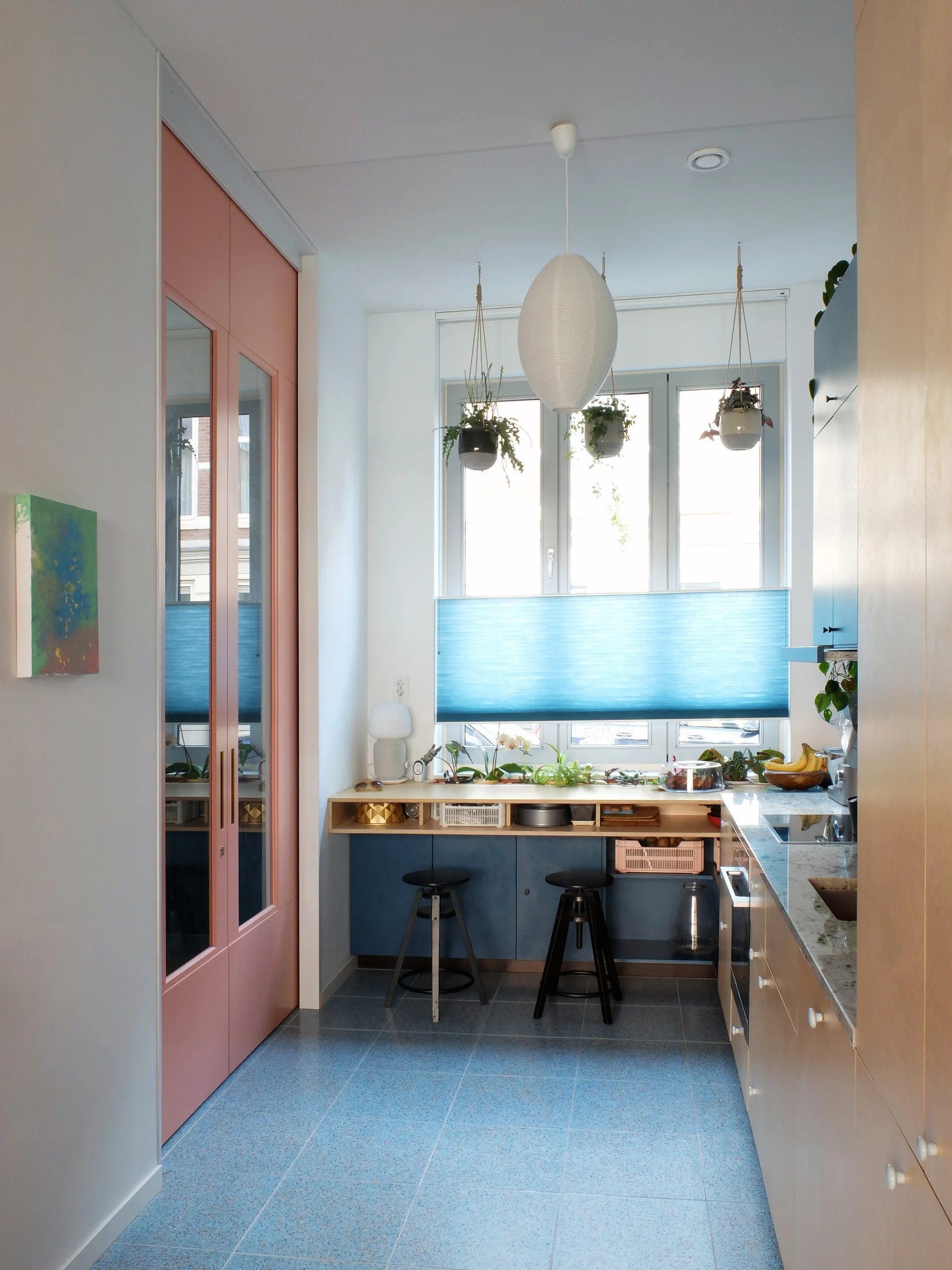 A colorful galley kitchen features blue speckled floor tiles, a pink closet door, light wood cabinets, and a breakfast bar beneath a sunny window.