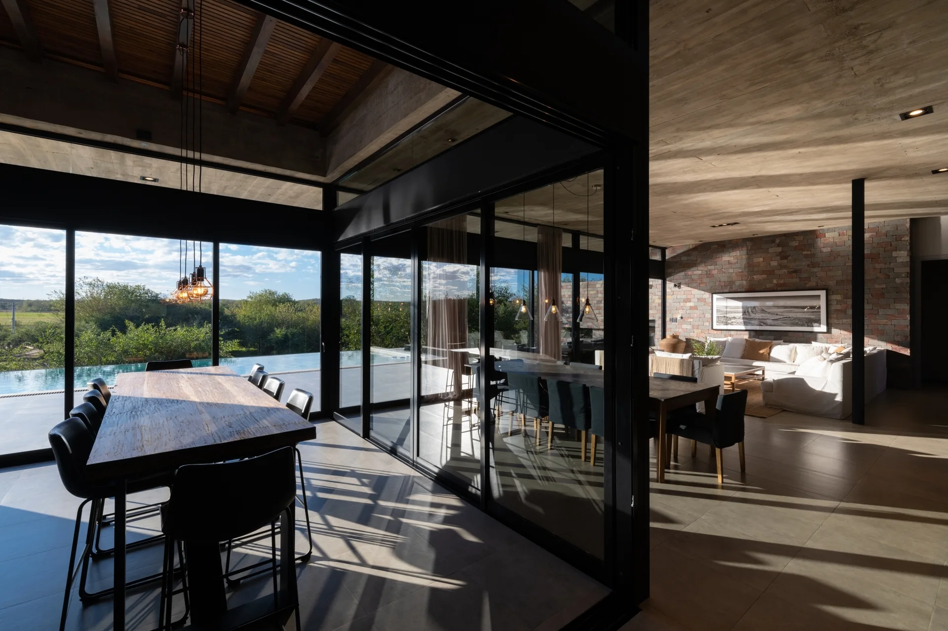 A modern open-concept dining room with a long wood table, large sliding glass partition walls, and a living area with a stone brick wall.