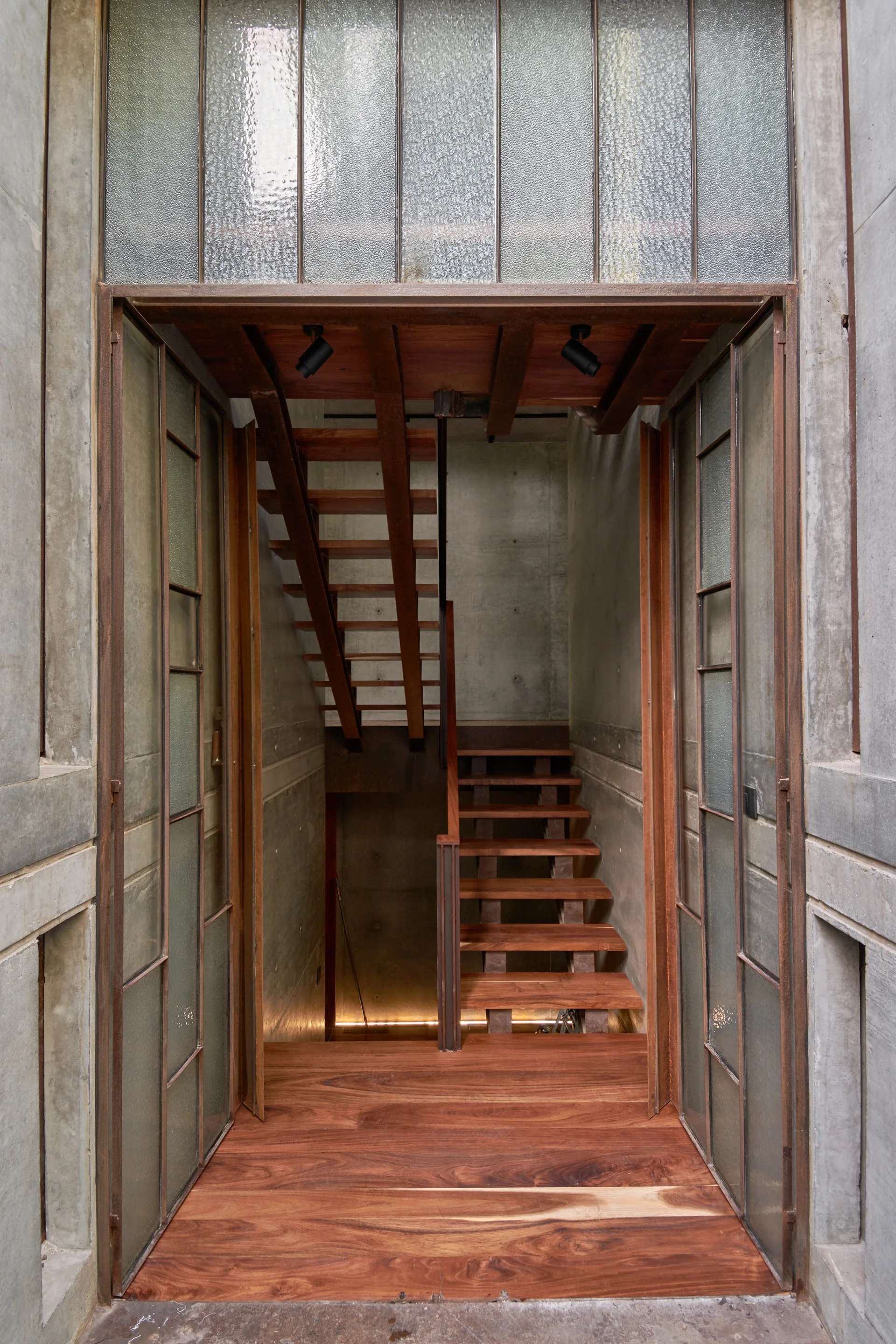 A wooden staircase climbs past raw concrete walls behind an industrial metal doorway with textured glass panels.