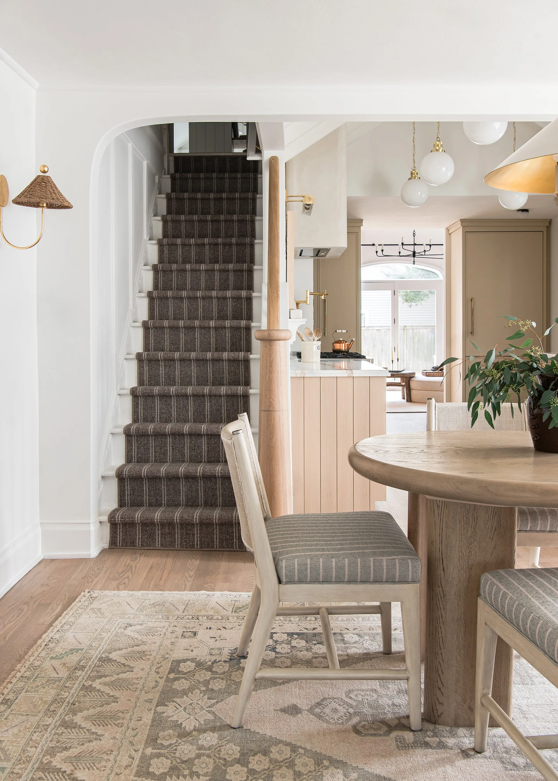 A dining area features a round wood table and cane-back chairs next to an arched entryway revealing a staircase with a brown plaid carpet runner.
