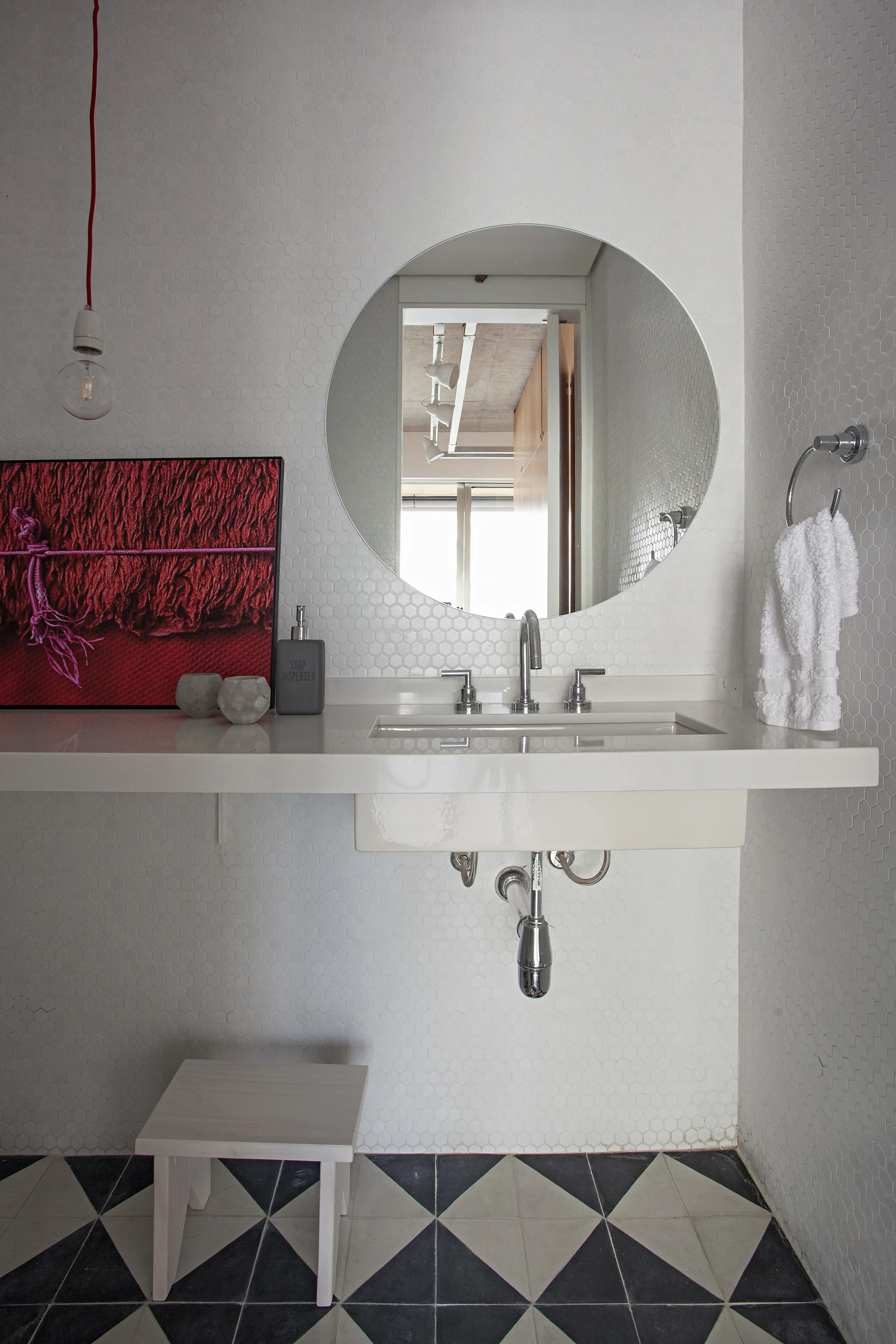 A minimalist bathroom with a wall-mounted sink, a round mirror, small white hexagonal wall tiles, and a bold black and white geometric floor.