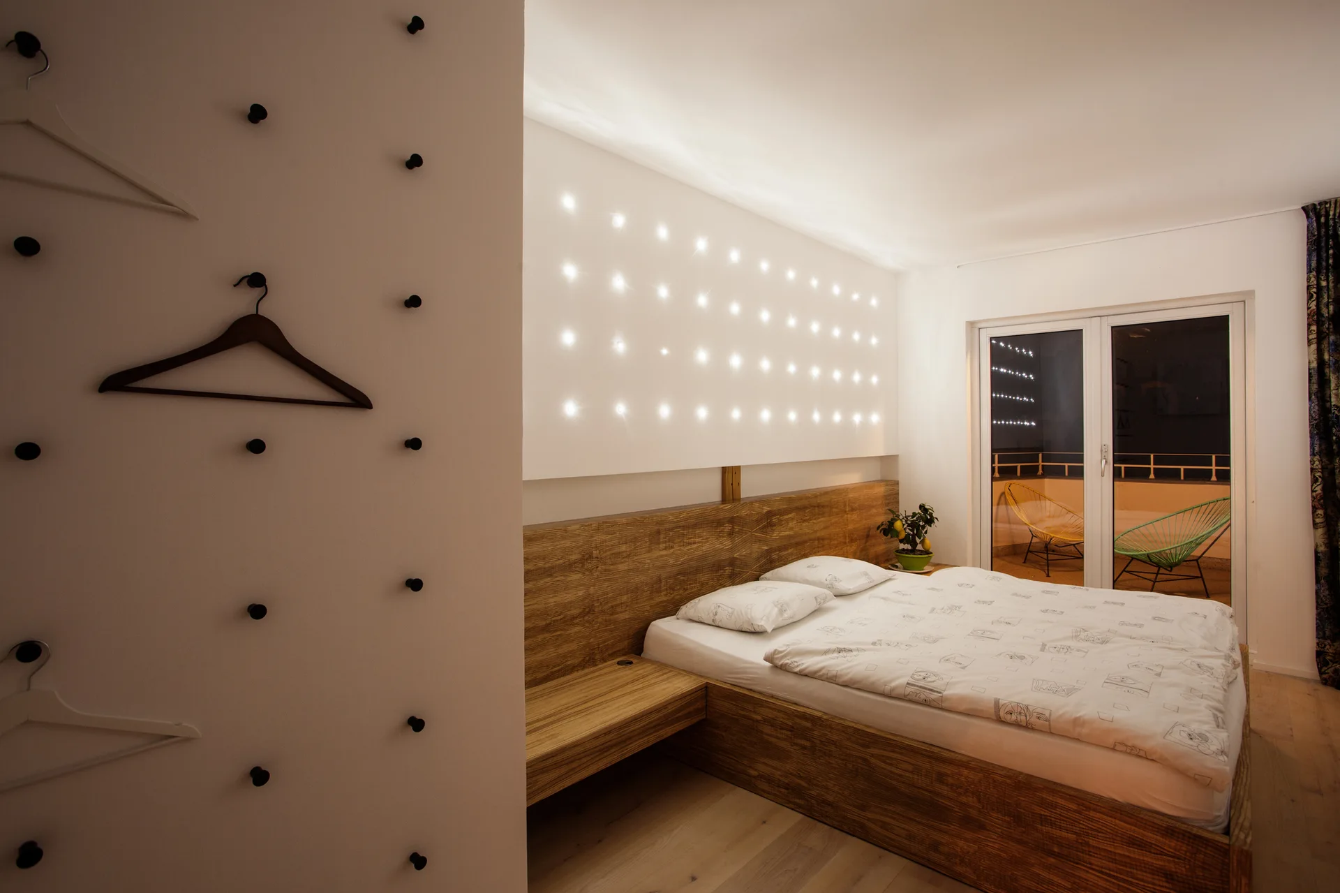 A modern bedroom features a wooden platform bed, an illuminated wall panel, and a white wall fitted with black hanging pegs.