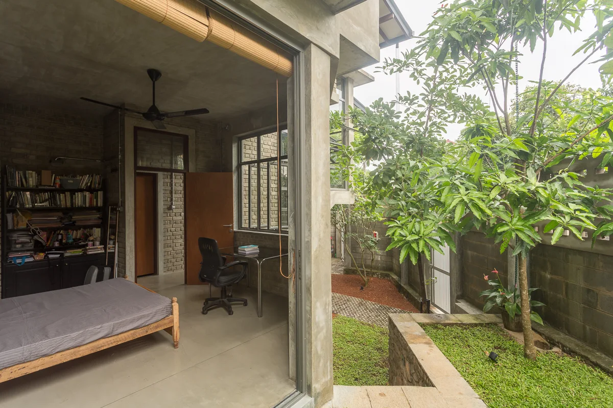 A minimalist grey concrete bedroom featuring a wooden bed, a glass desk, and a large sliding glass door that opens onto a green courtyard.