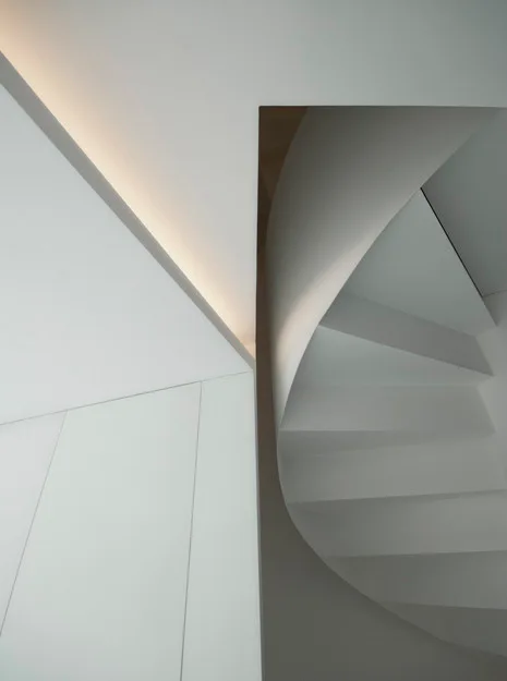 An architectural view of a minimalist white curved staircase illuminated by soft recessed cove lighting along the wall and ceiling.