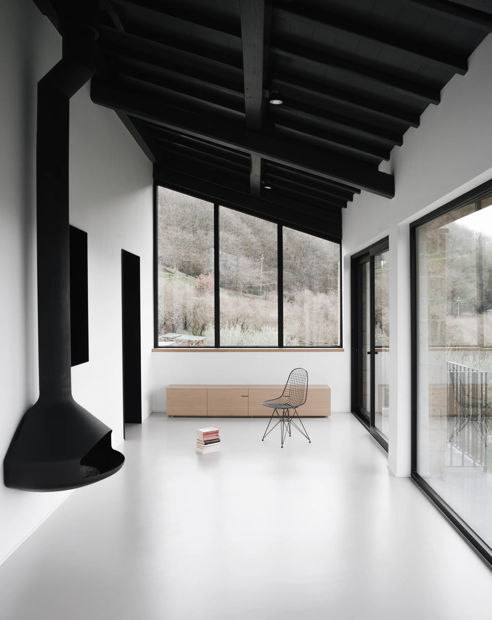 A minimalist white living space features a black suspended fireplace, a wire chair, a low wooden sideboard, and a dark beamed vaulted ceiling.