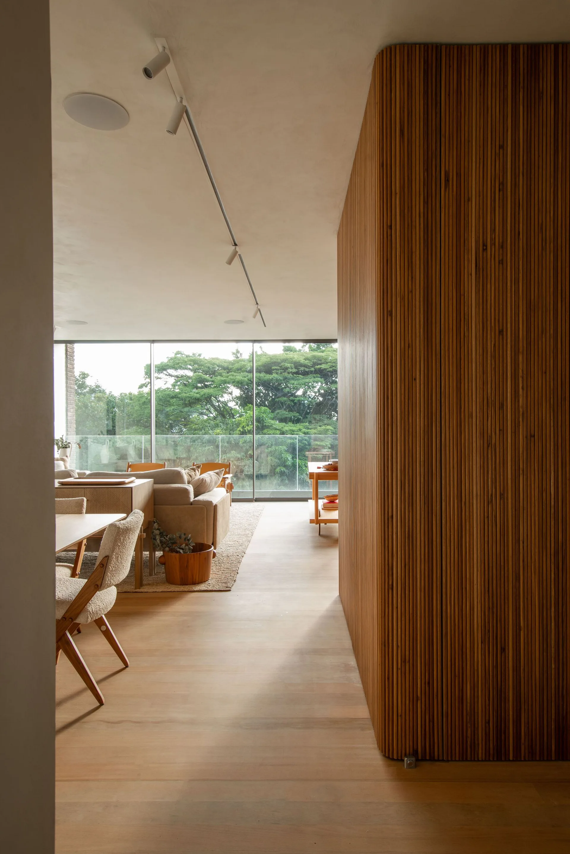 A modern open-plan living and dining area features a curved slatted wood wall partition, light wood floors, and a large window looking out to trees.