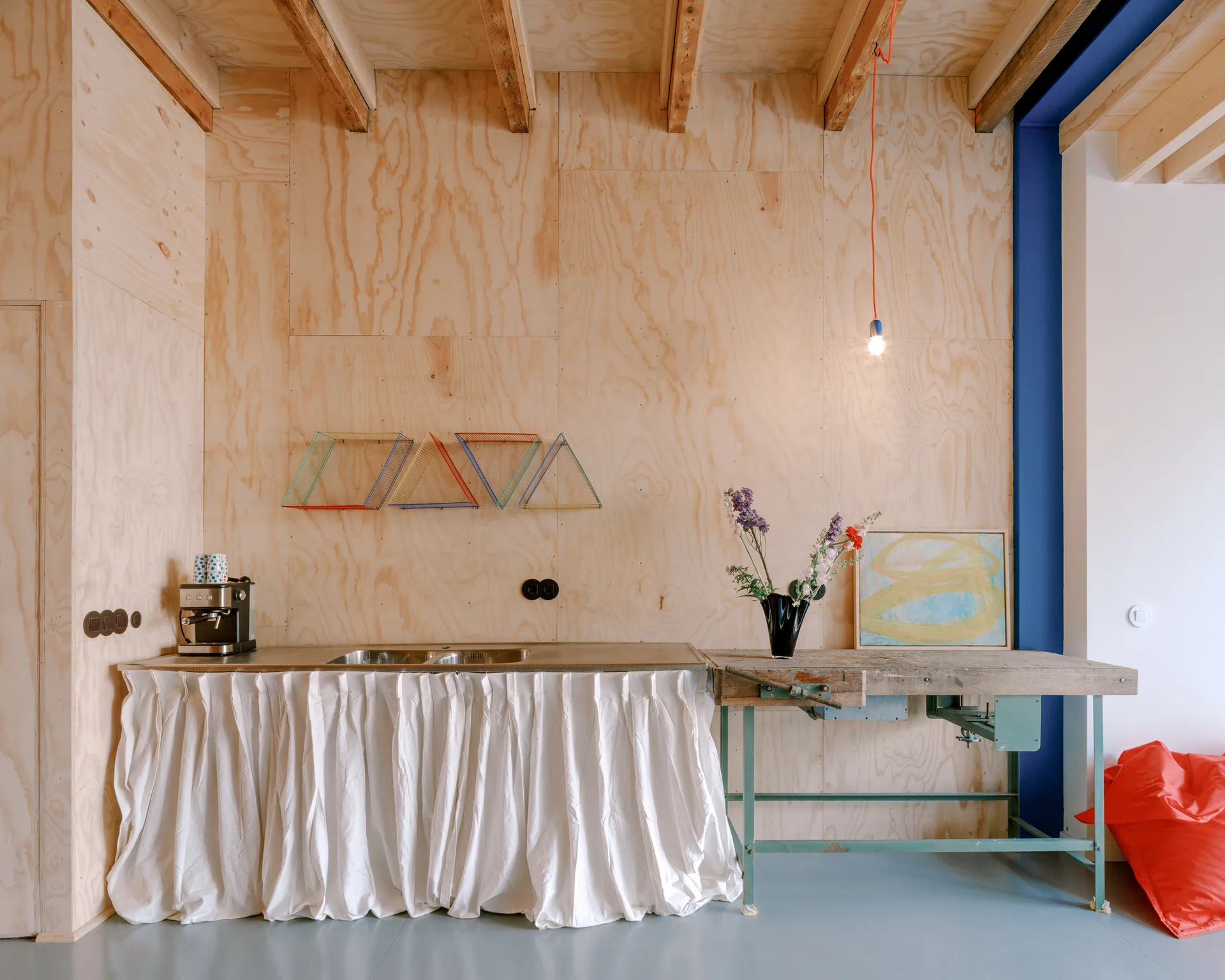 A utilitarian kitchen space with warm plywood walls, a white fabric skirt under the sink, and a rustic wooden workbench used as a counter.