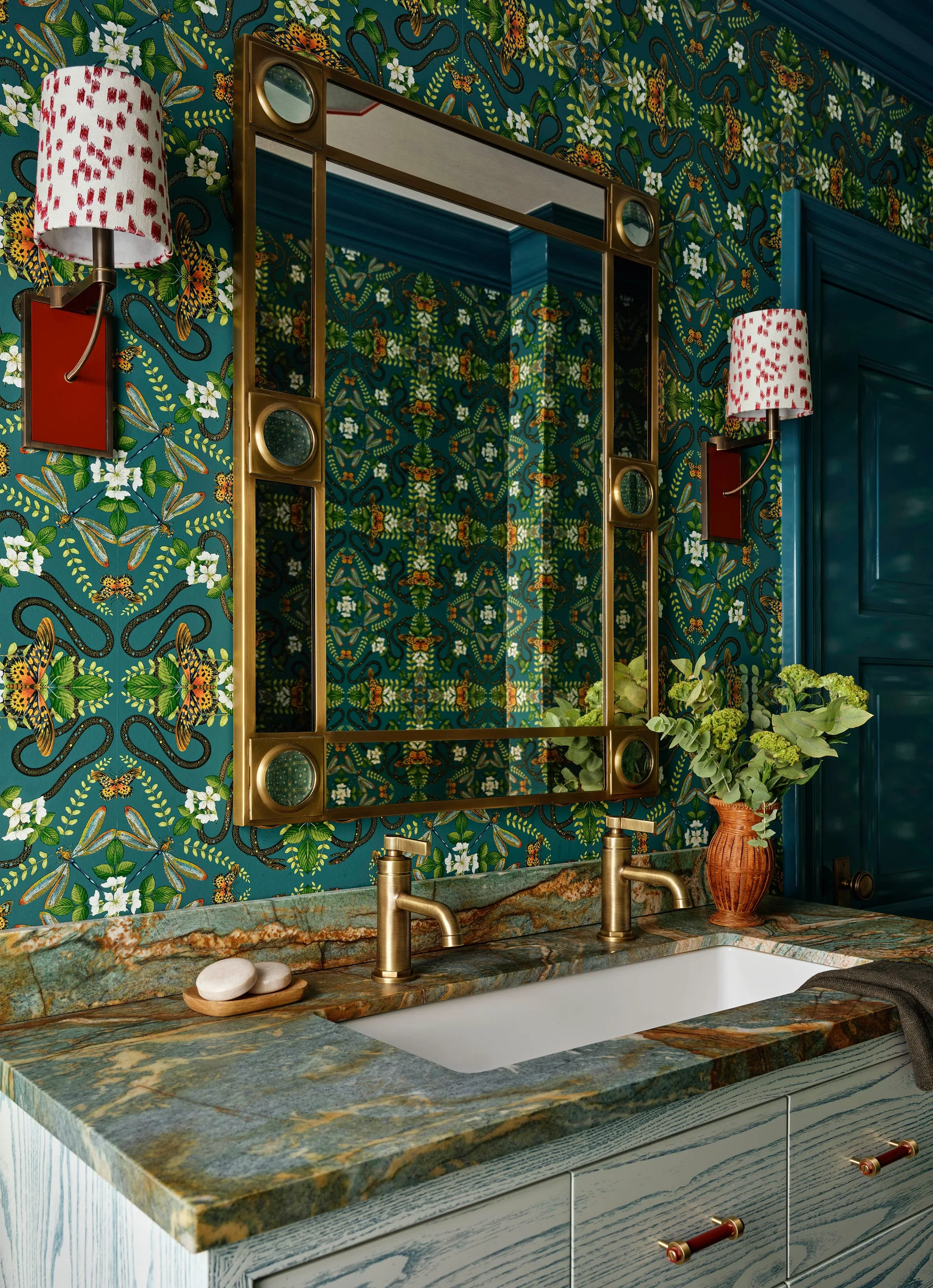 A bathroom vanity featuring a heavily veined green and brown stone countertop with brass faucets set against dark green botanical wallpaper.