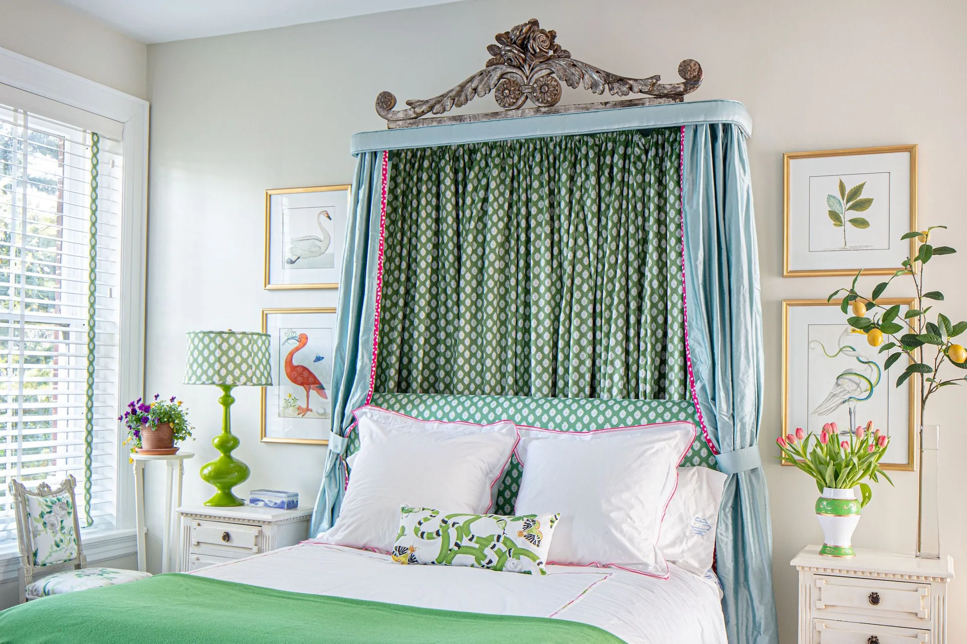 A traditional bedroom features a canopy bed with a carved wood pediment, light blue drapes, green patterned fabric, and white nightstands.