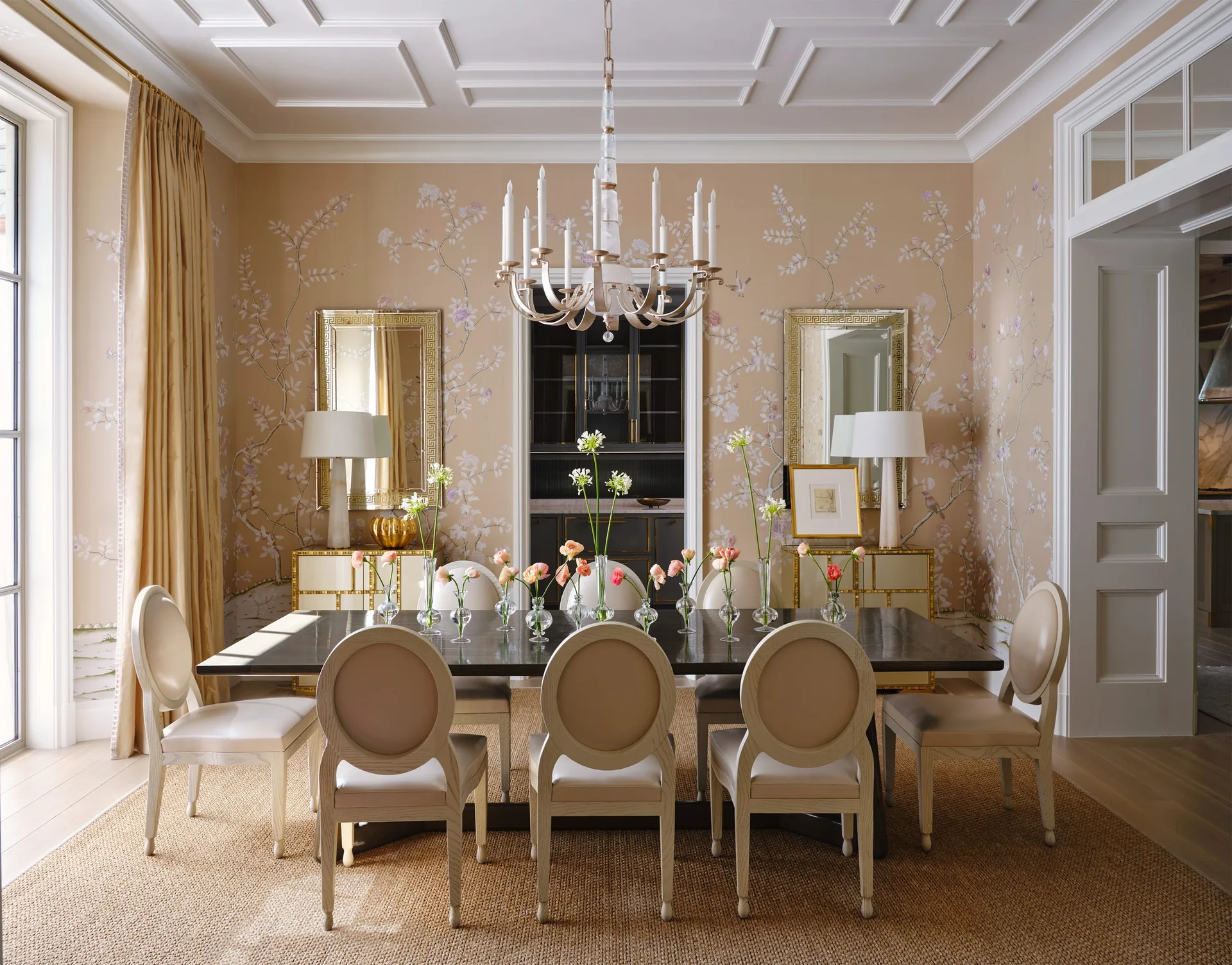 A formal dining room features a dark wood table with small flower vases, oval-back chairs, beige floral wallpaper, and twin mirrors flanking a doorway.