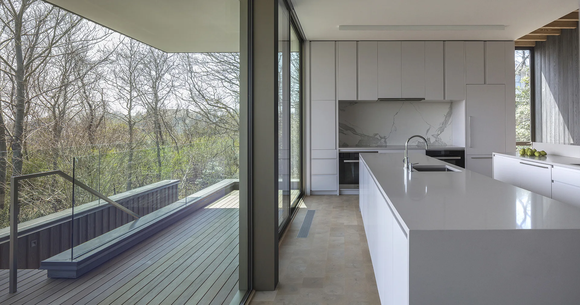 A minimalist white kitchen features a long island with a sink, flat-panel cabinets, and a sliding glass door opening to a wooden deck.