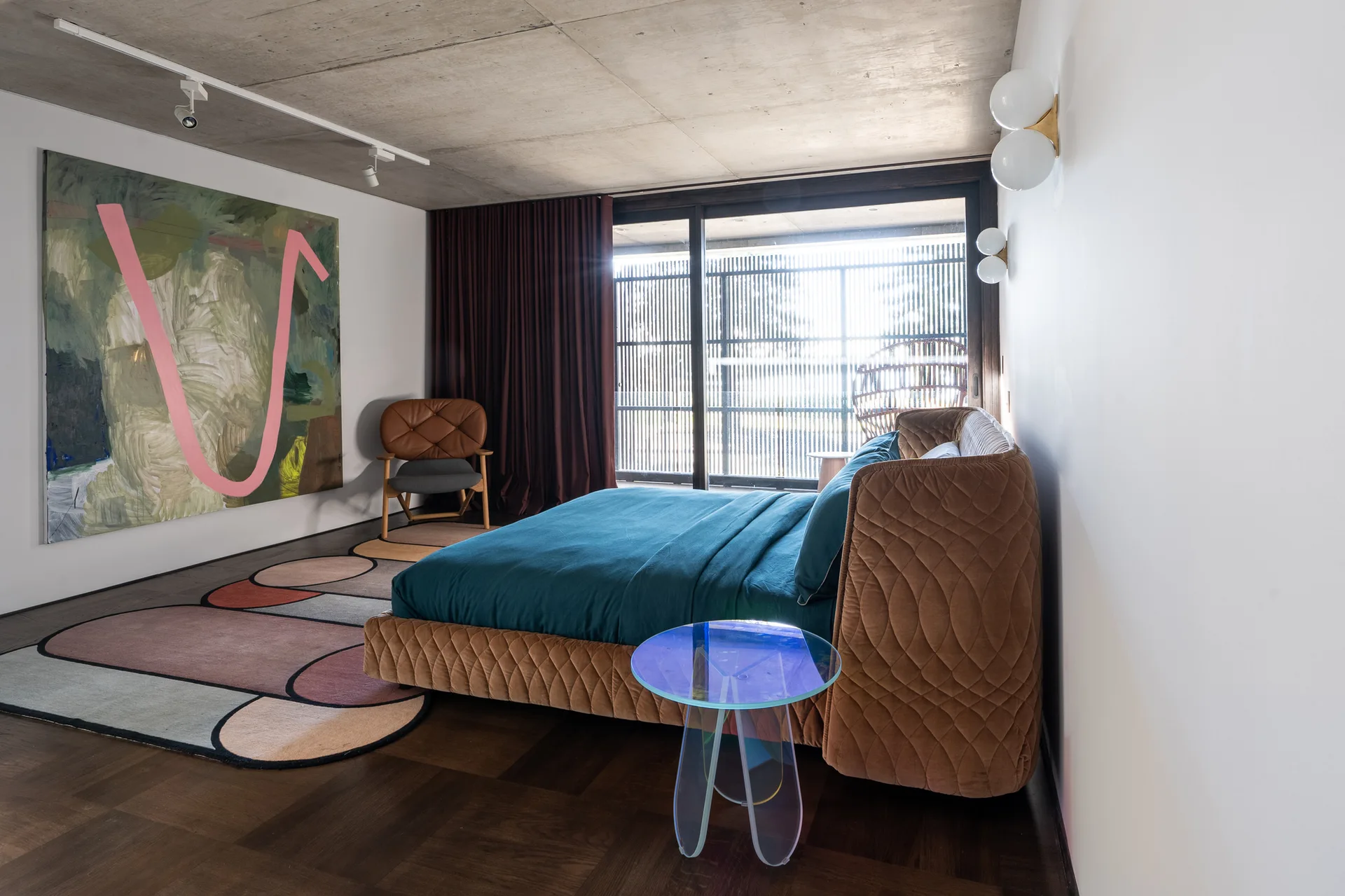 A contemporary bedroom featuring a tan quilted bed, an iridescent side table, and a large abstract painting beneath an exposed concrete ceiling.