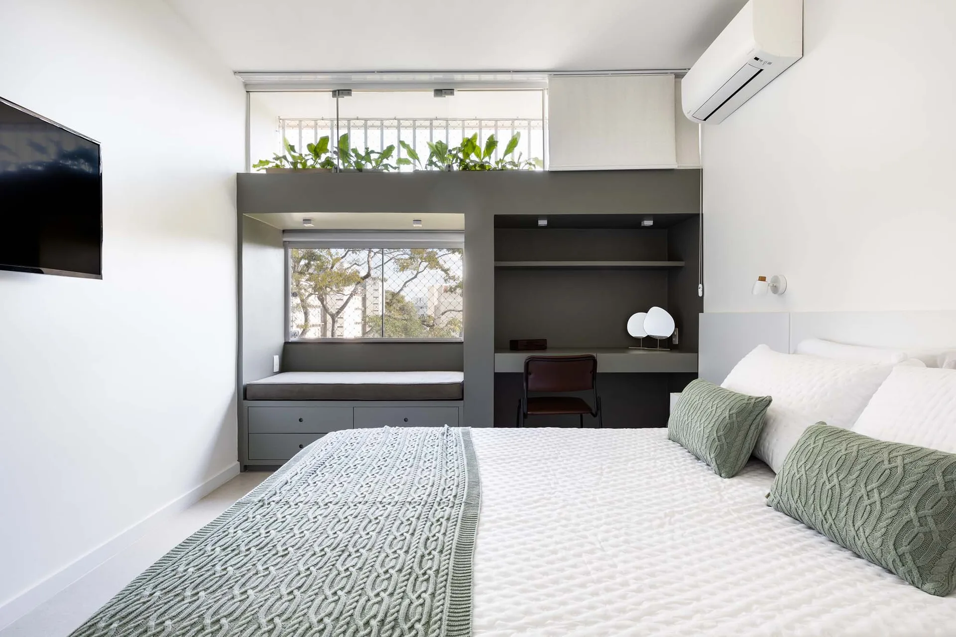A modern white bedroom features a gray built-in unit containing a workspace, window seat, and top planter shelf.