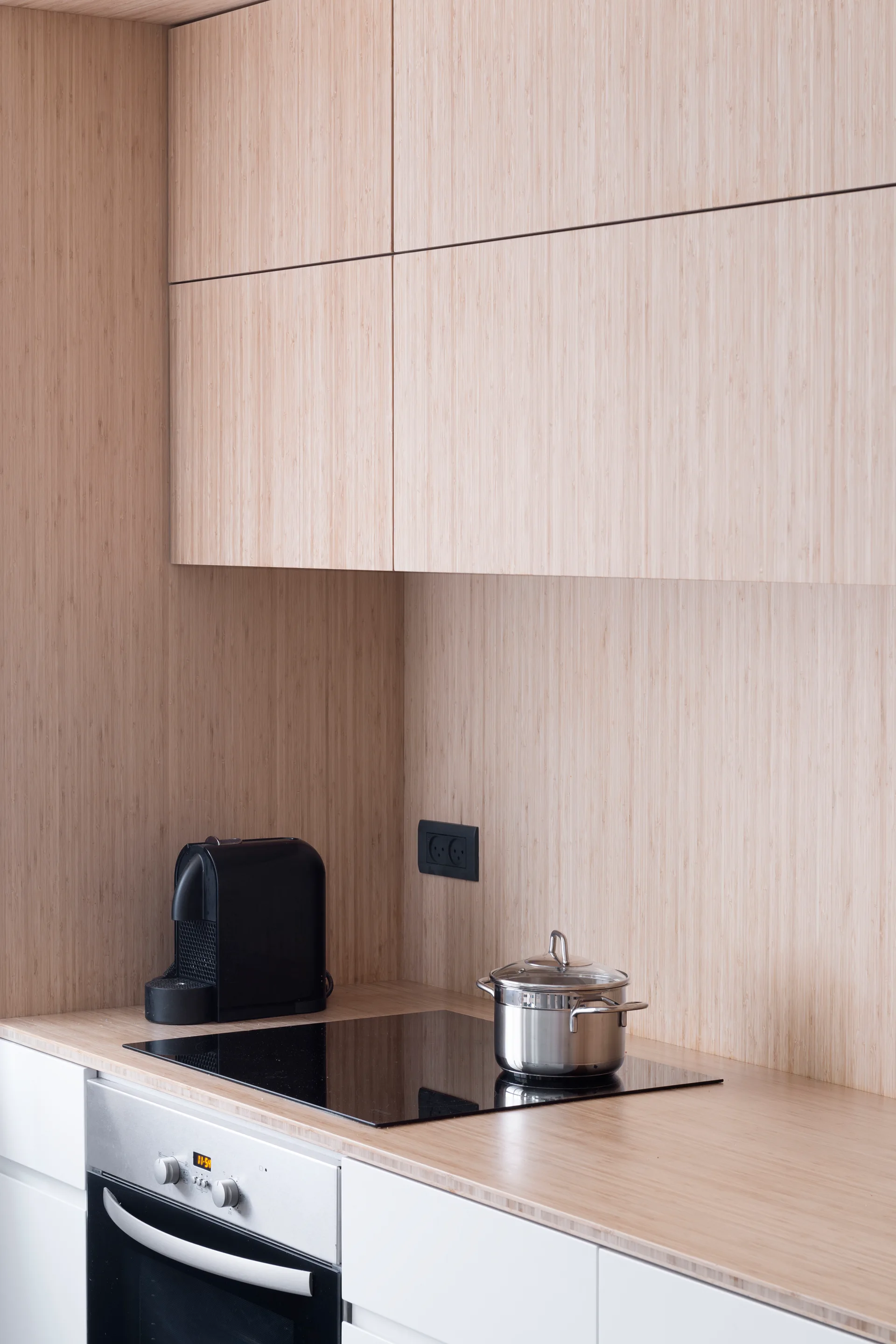 Minimalist kitchen corner featuring light wood cabinets and matching wood backsplash, a black cooktop, and a stainless steel oven.
