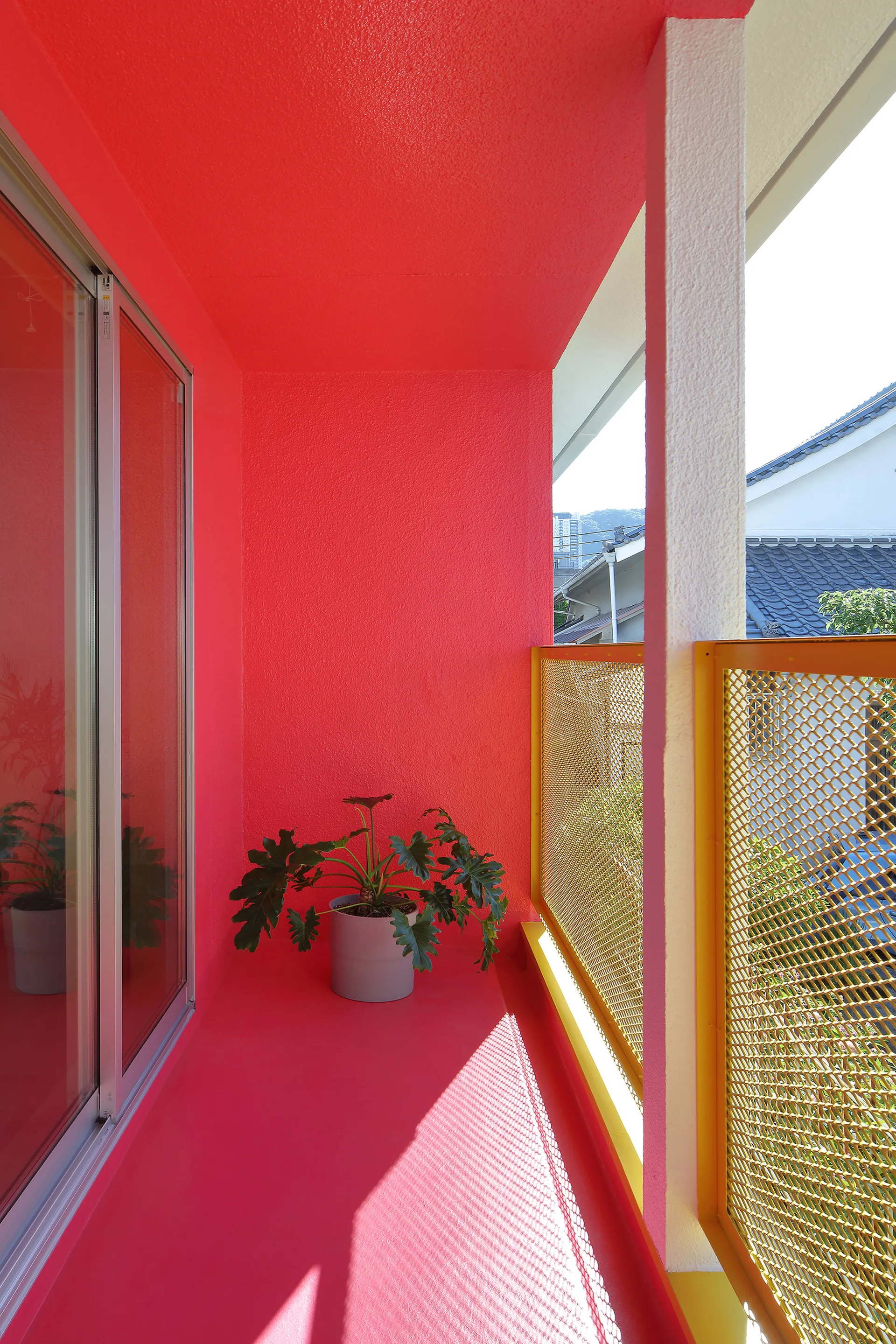 A narrow balcony painted entirely in red features a bright yellow metal mesh railing and a green potted plant in a gray planter.