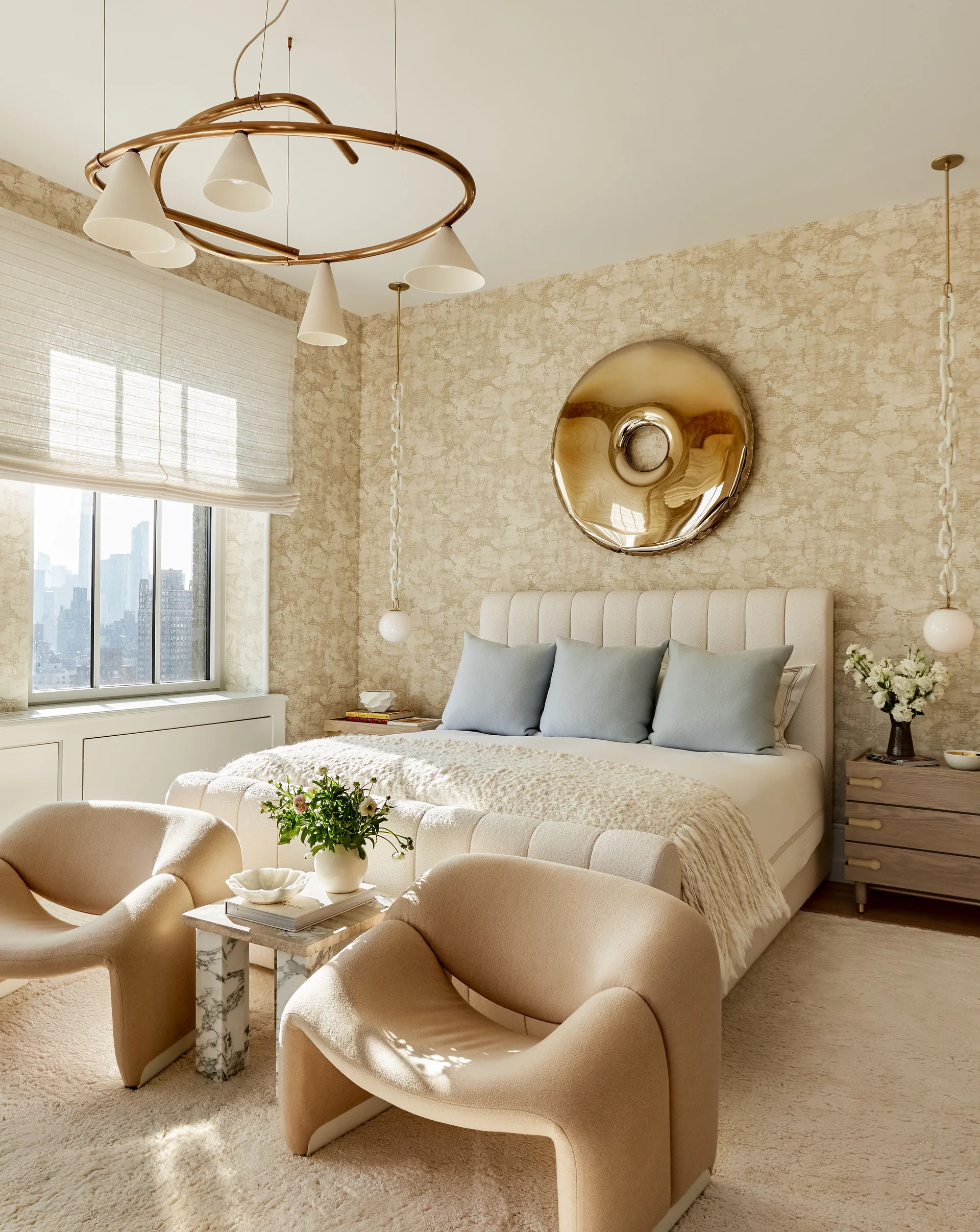 A beige contemporary bedroom features a cream upholstered bed, two curved armchairs, a gold ring chandelier, and a circular metallic wall sculpture.