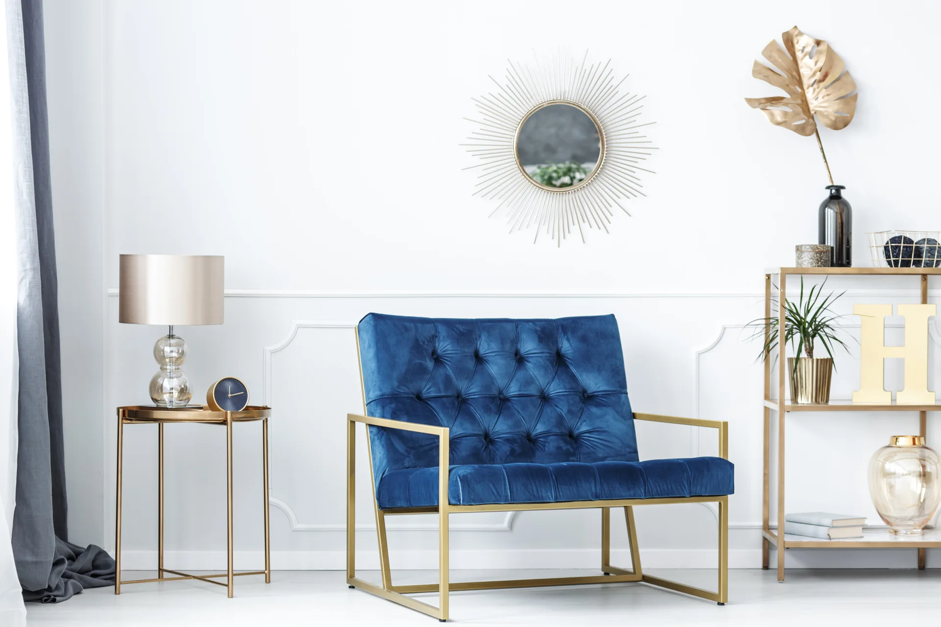 A tufted blue velvet armchair sits between a gold side table and a gold shelving unit against a white paneled wall with a sunburst mirror.