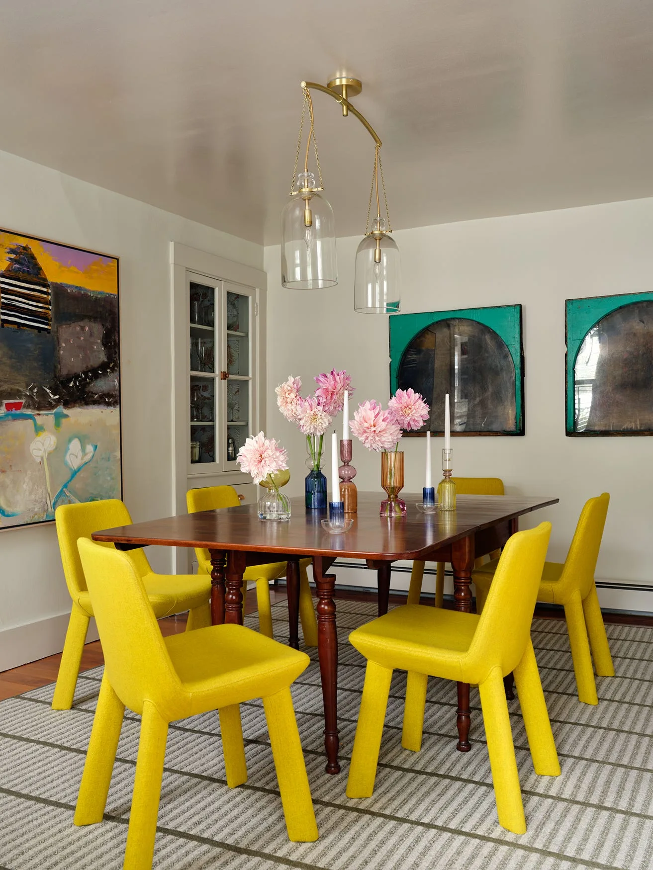 An eclectic dining room with a dark wooden table, six bright yellow upholstered chairs, colorful glass vases, and a gold glass pendant light.
