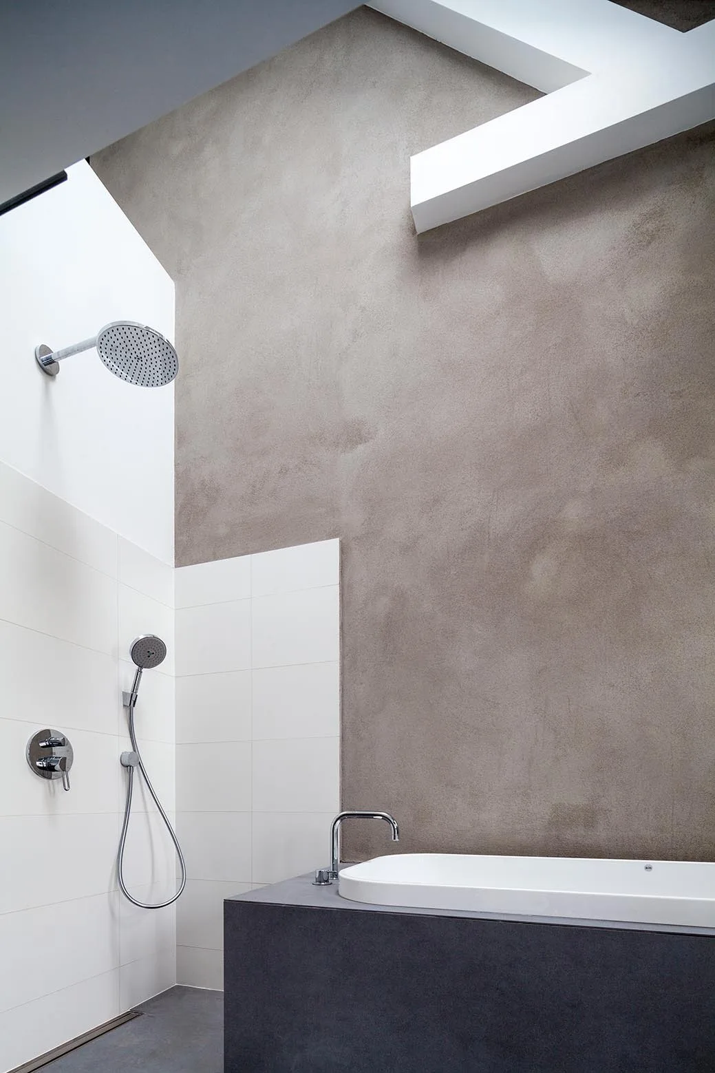 A modern bathroom showing a white bathtub with a dark grey base set against a textured grey plaster wall next to a white tiled shower area.