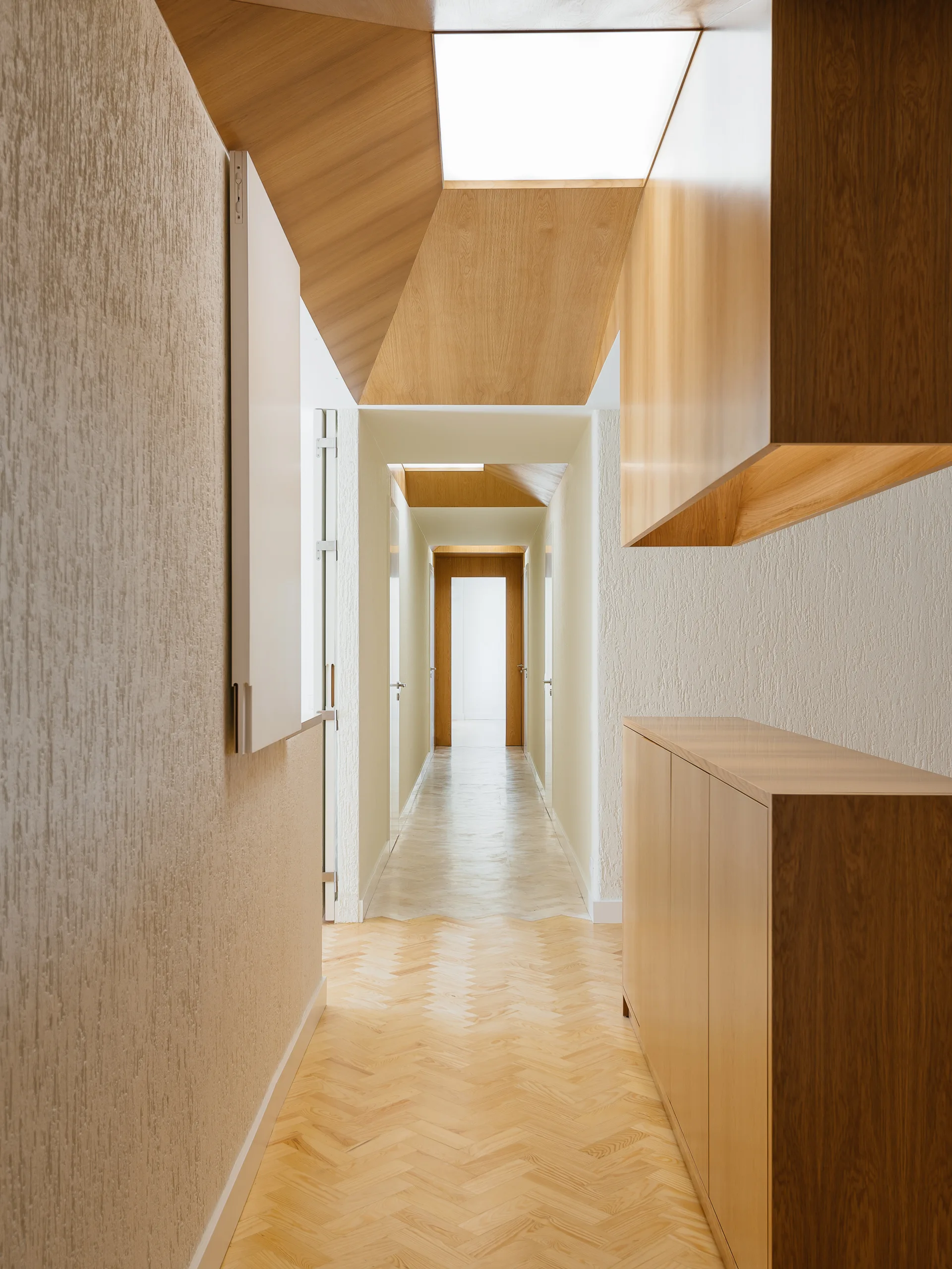 A long minimalist hallway features light wood herringbone flooring, textured off-white walls, and custom wood-paneled ceiling details.