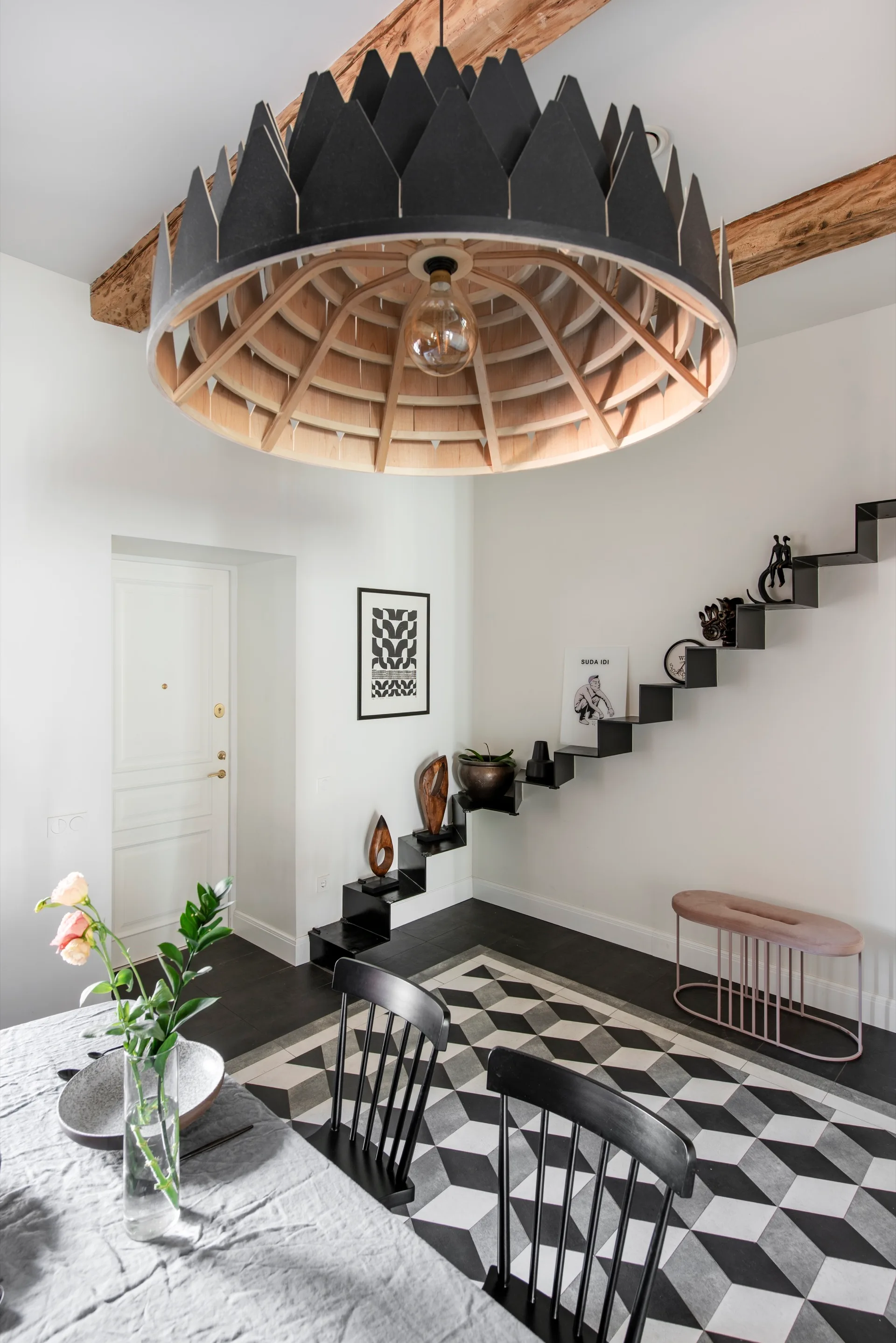 A modern dining room features a large black wooden dome pendant light, a black-and-white geometric tiled floor, and a black floating staircase.