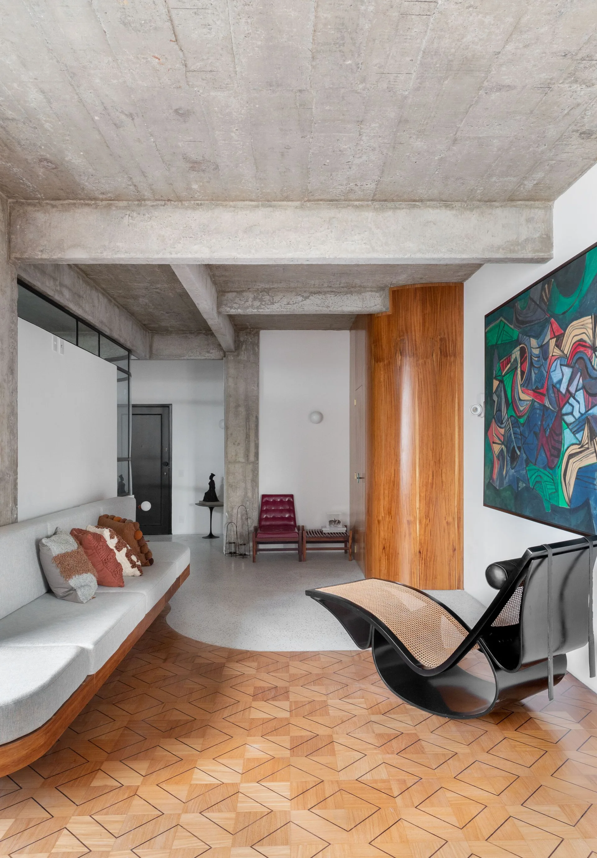 A living room features an exposed concrete ceiling, geometric parquet flooring, a curved wood wall panel, a gray sofa, and a black lounge chair.