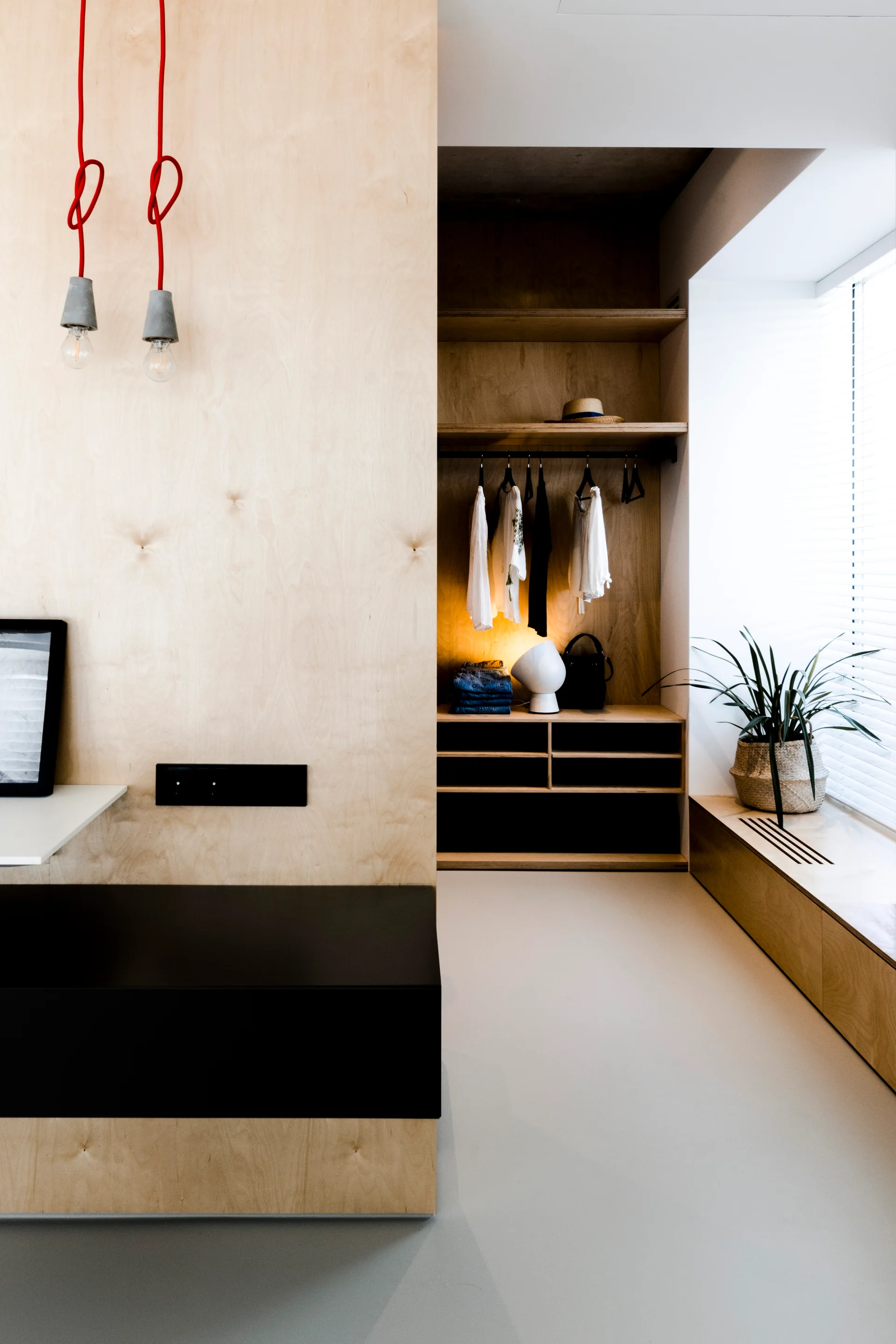 A minimalist dressing area features a light plywood closet system, an integrated wooden window bench, and two hanging lightbulbs with red cords.