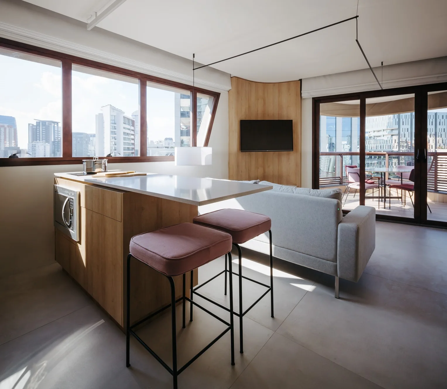 A modern open-concept apartment interior features a wood-clad kitchen island with two pink bar stools, a gray sofa, and a curved wood accent wall.