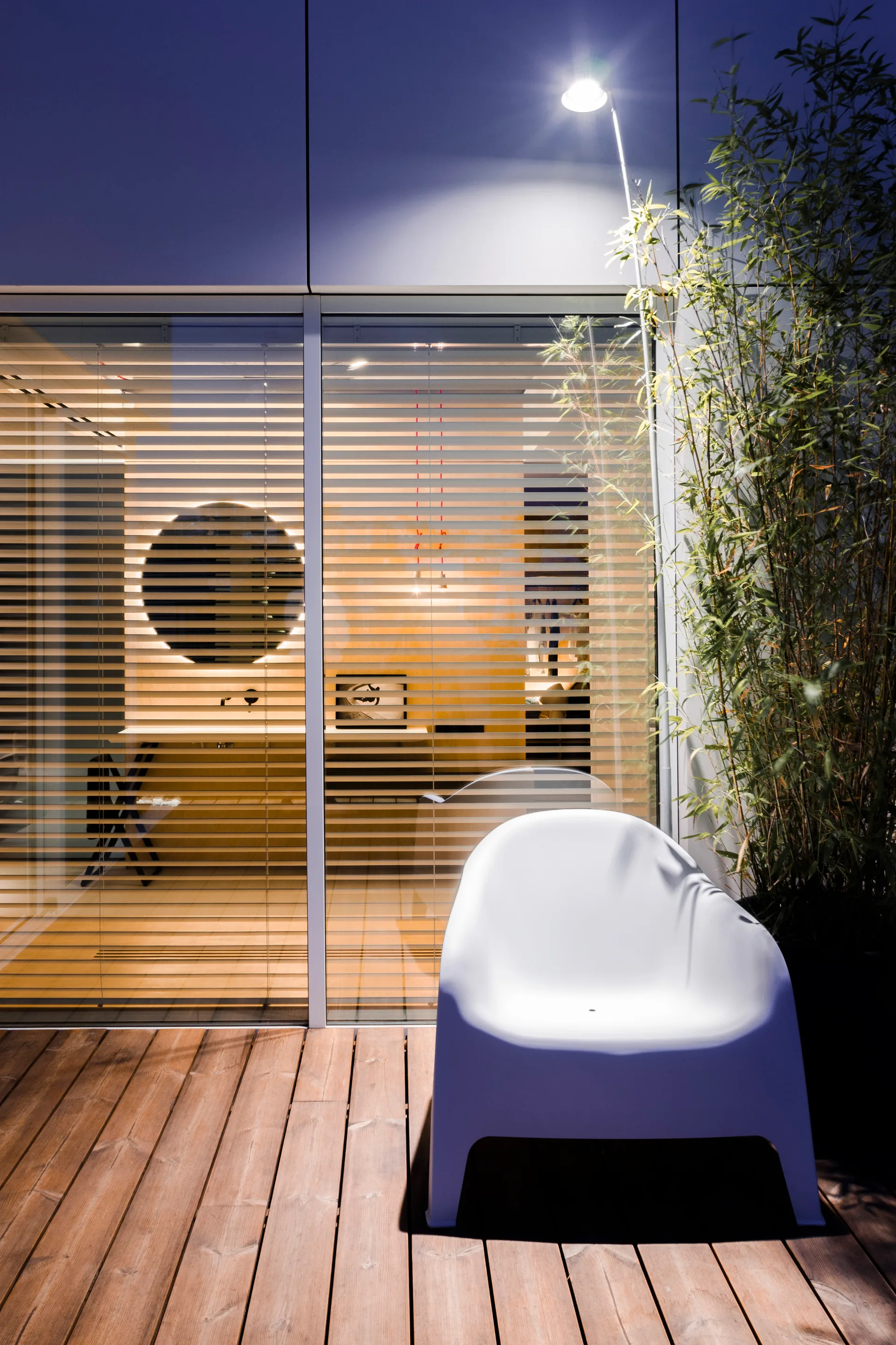 A modern wooden patio deck at night features a white sculptural armchair, a tall potted bamboo plant, and horizontal window blinds showing a lit interior.