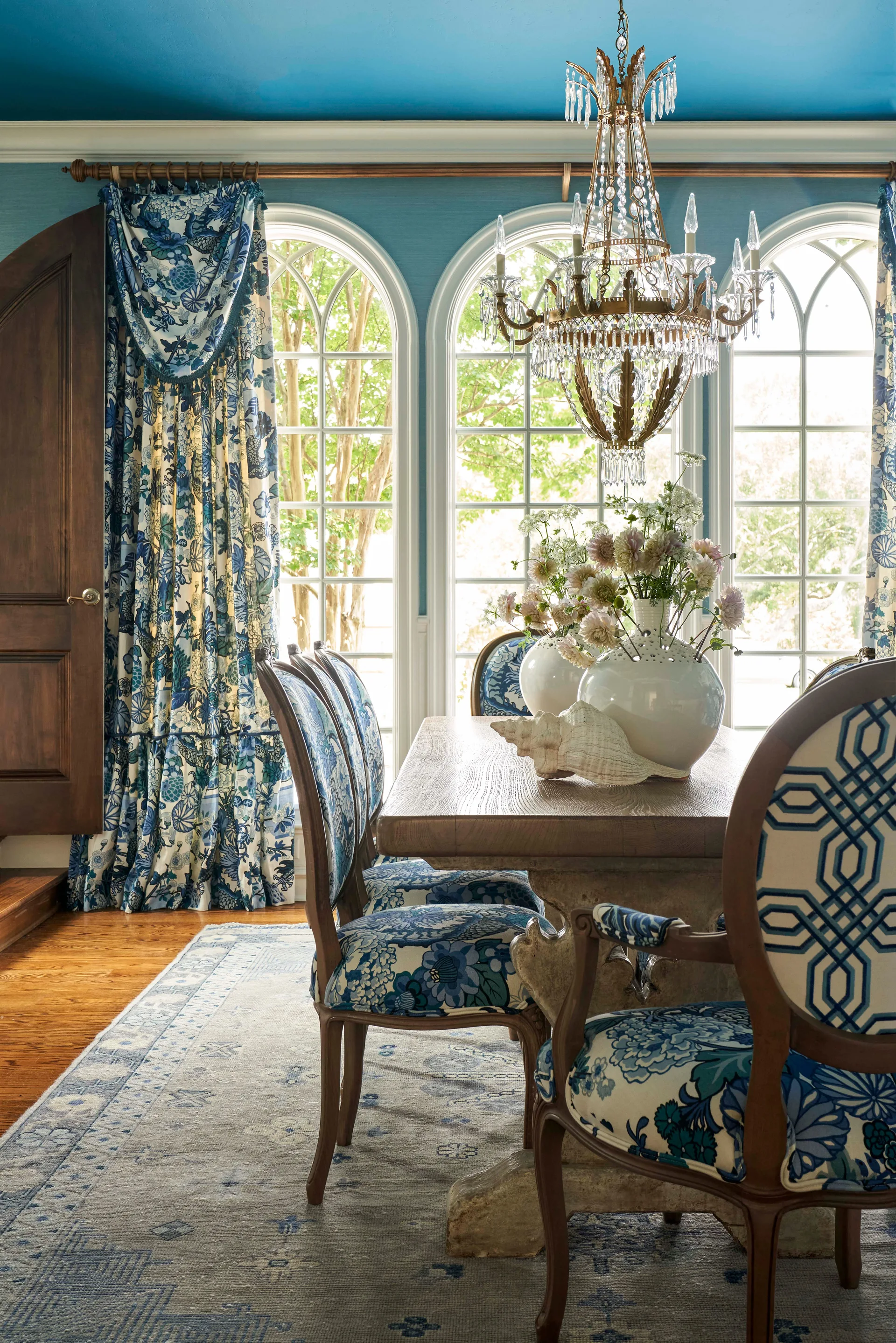 A dining table with blue floral upholstered chairs sits beneath a crystal chandelier in a room with blue walls, arched windows, and patterned curtains.
