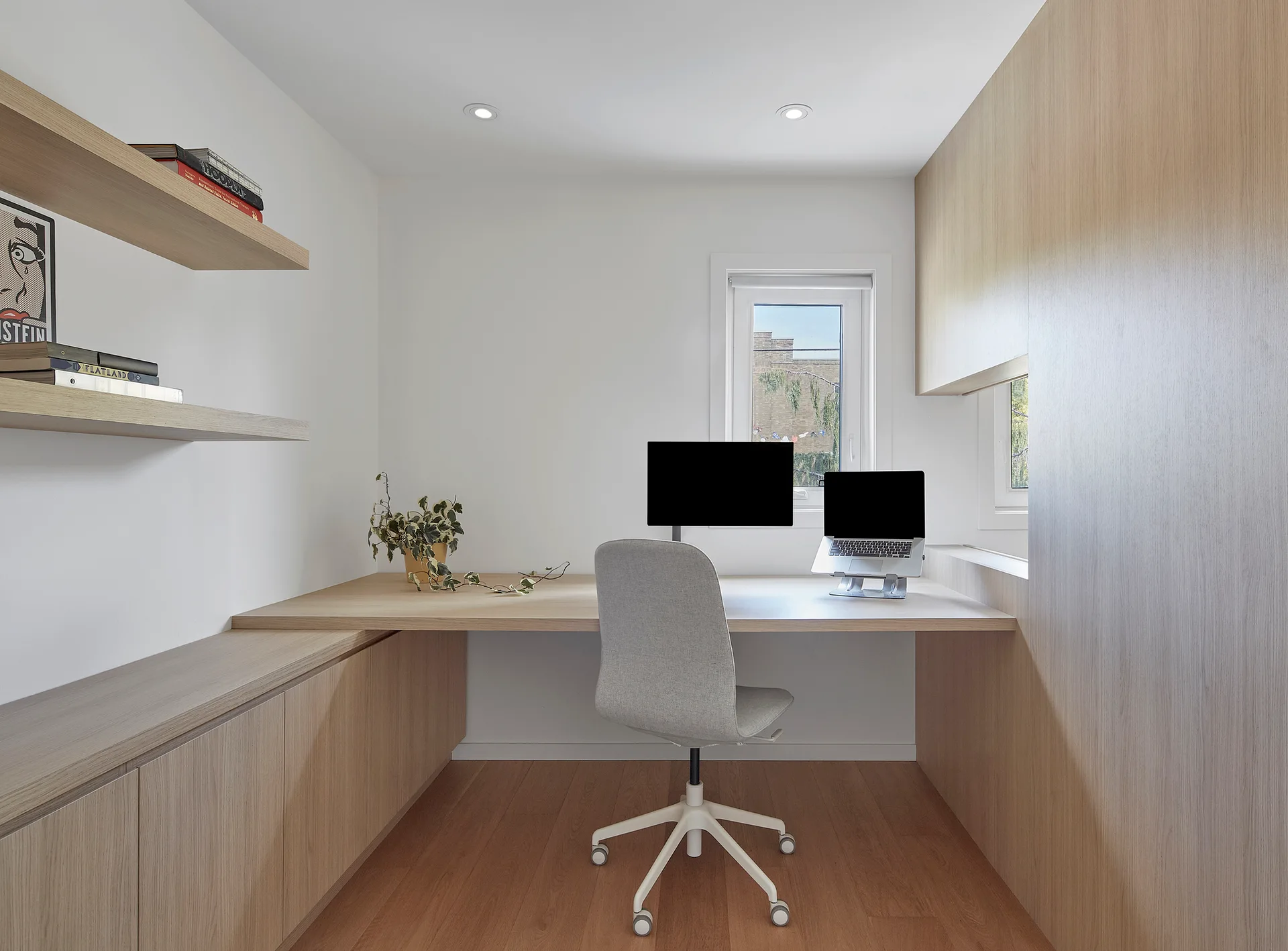 A compact home office features a built-in light wood desk, matching cabinets, a grey swivel chair, and floating shelves against white walls.