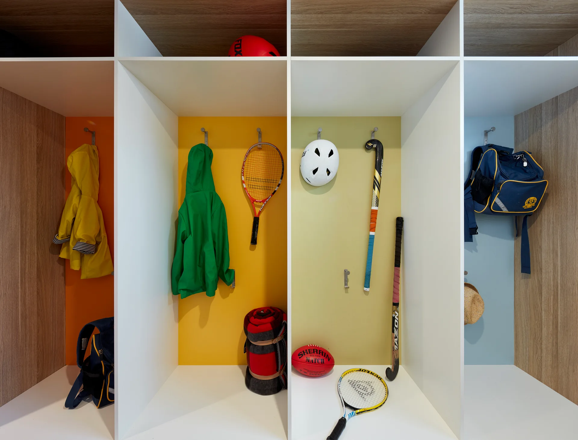 Four vertical mudroom cubbies feature color-coded back panels in orange, yellow, green, and blue, with white dividers and hanging sports gear.