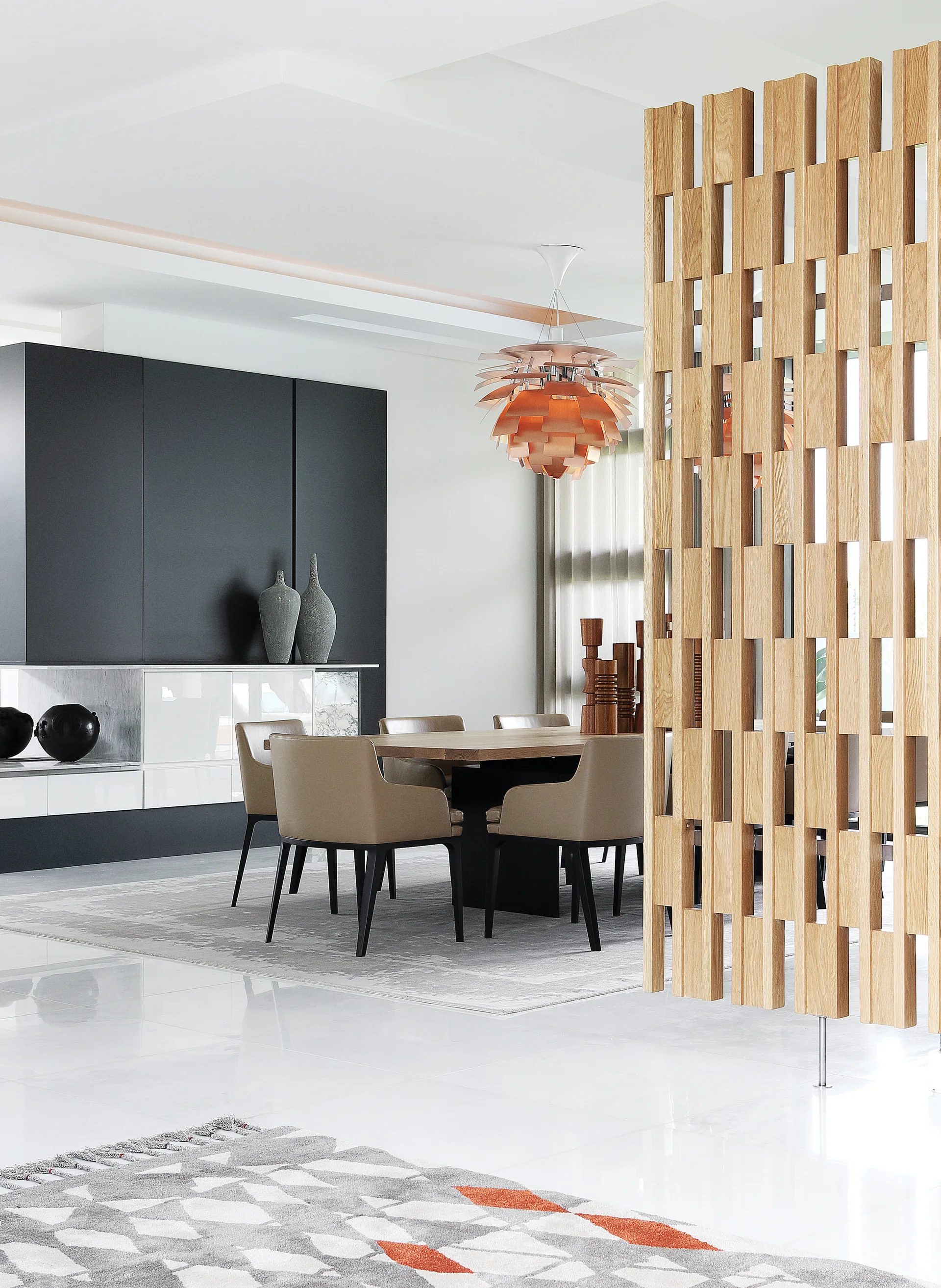 A modern dining room features a wooden table, tan chairs, a copper pendant light, and a vertical wood slat partition on a white polished floor.