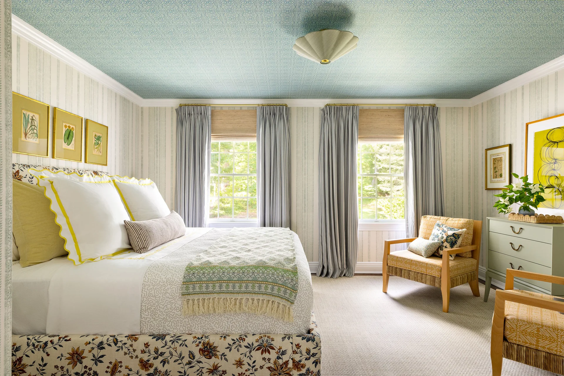 A well-lit bedroom featuring a bed with white and yellow bedding, striped wallpaper, a patterned blue ceiling, and twin windows with grey drapery.