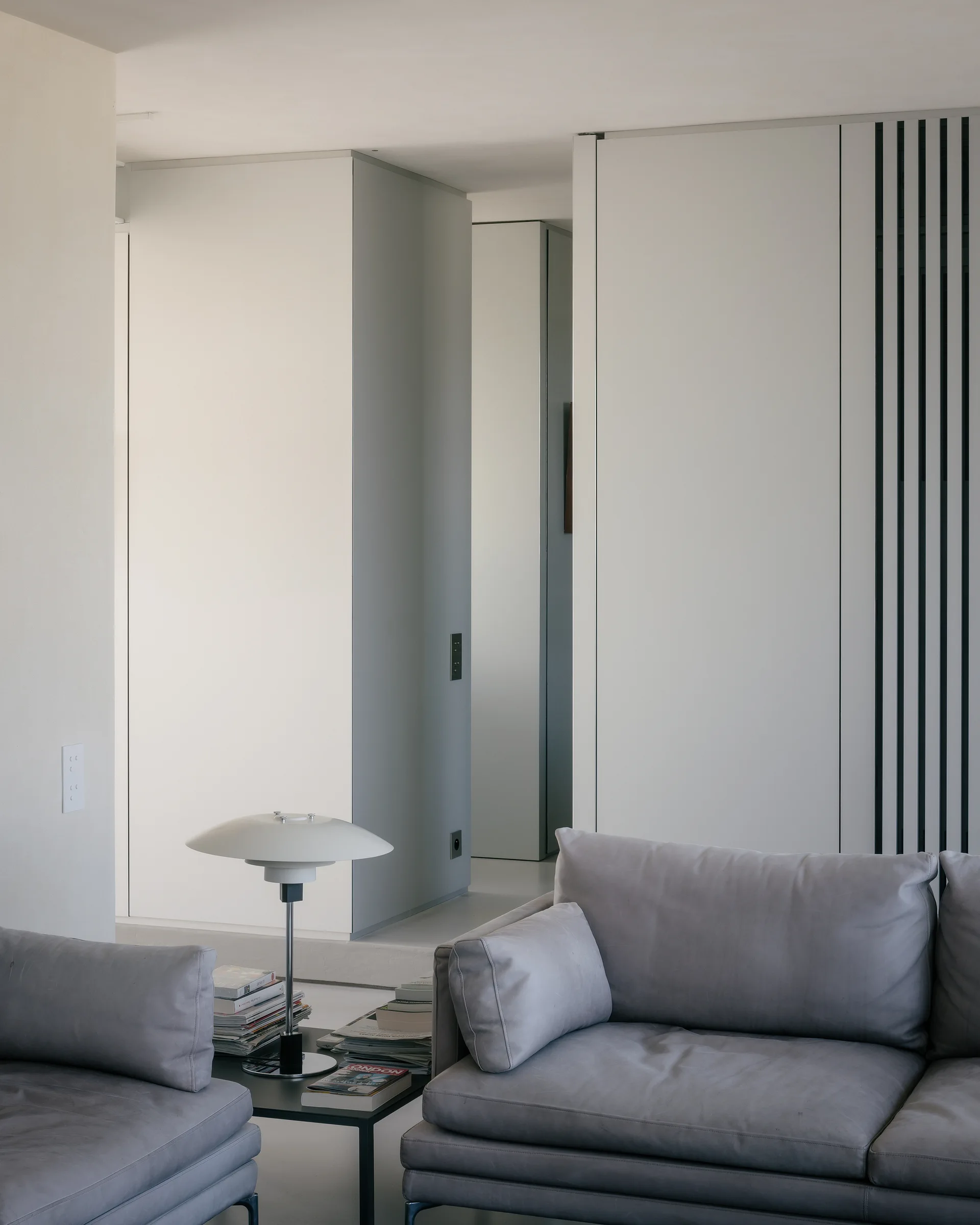 A minimalist living room features gray leather sofas flanking a metal side table with a mushroom-shaped lamp against white wall panels.